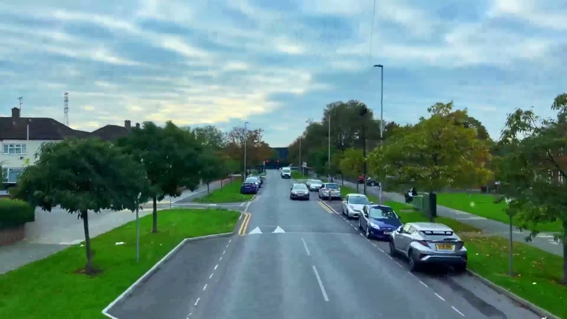 A residential street with parked and moving cars under a partly cloudy sky.
