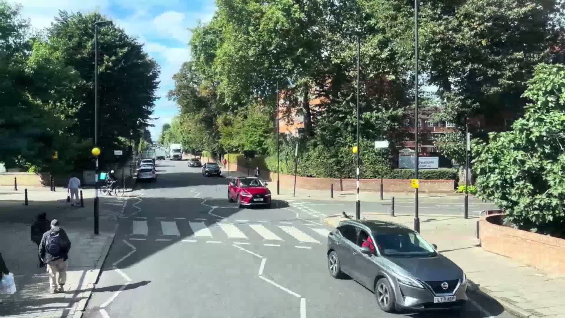 A sunny day shows a pedestrian crossing with several cars and pedestrians at an intersection lined with leafy trees.