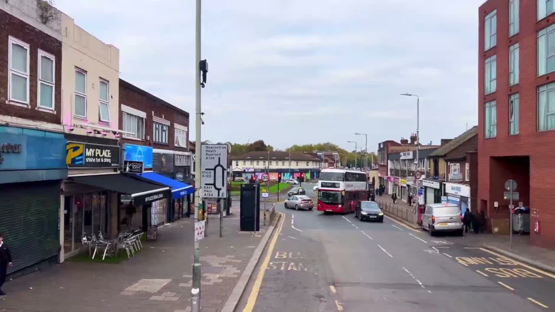 A street scene on an overcast day shows several shops and a double-decker bus at an intersection with cars and pedestrians.