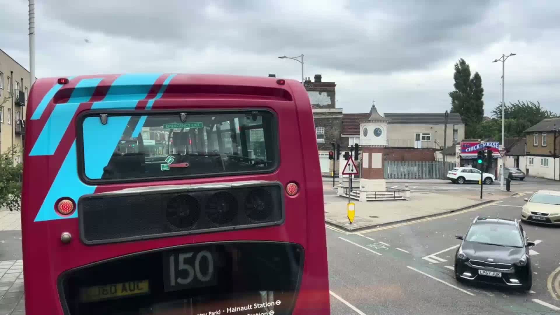 The rear of a double-decker bus, predominantly maroon with teal accents, is seen at a road intersection on an overcast day.