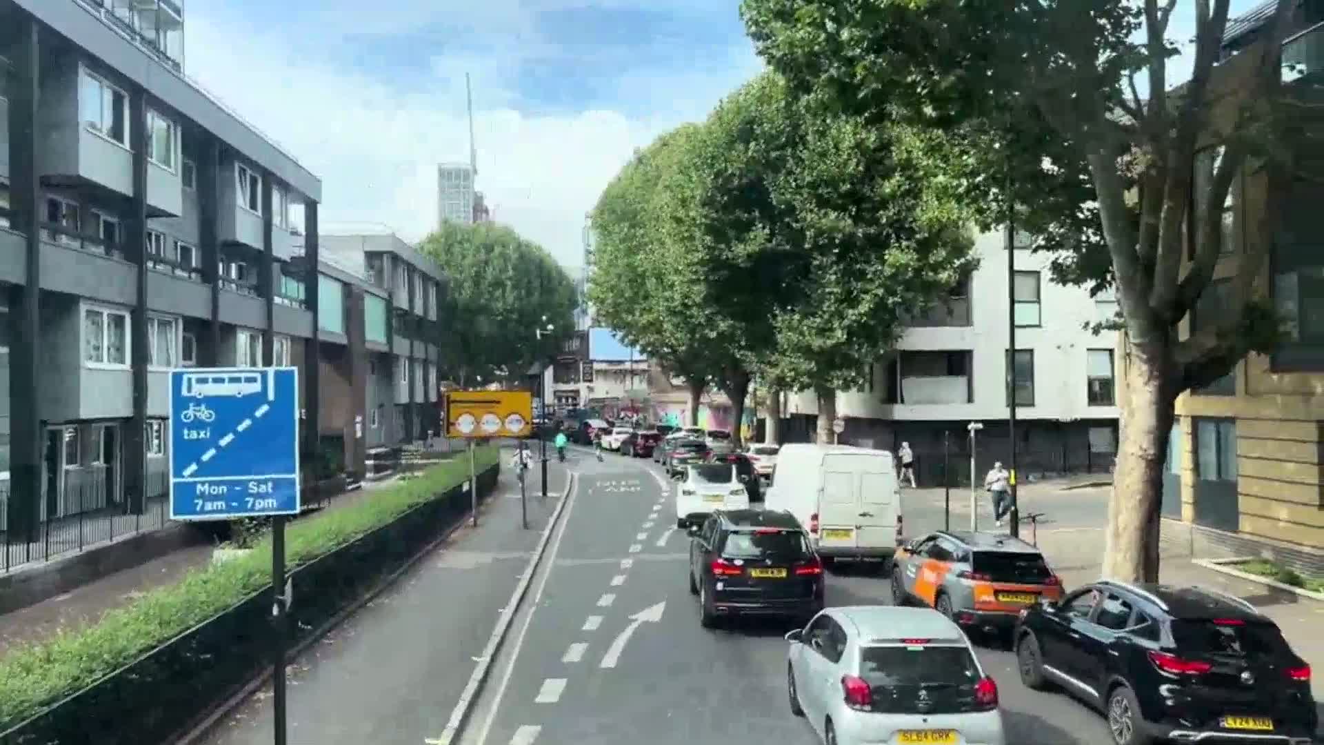 A sunny day shows a line of slow-moving cars and vans on a city street lined with apartment buildings and trees.
