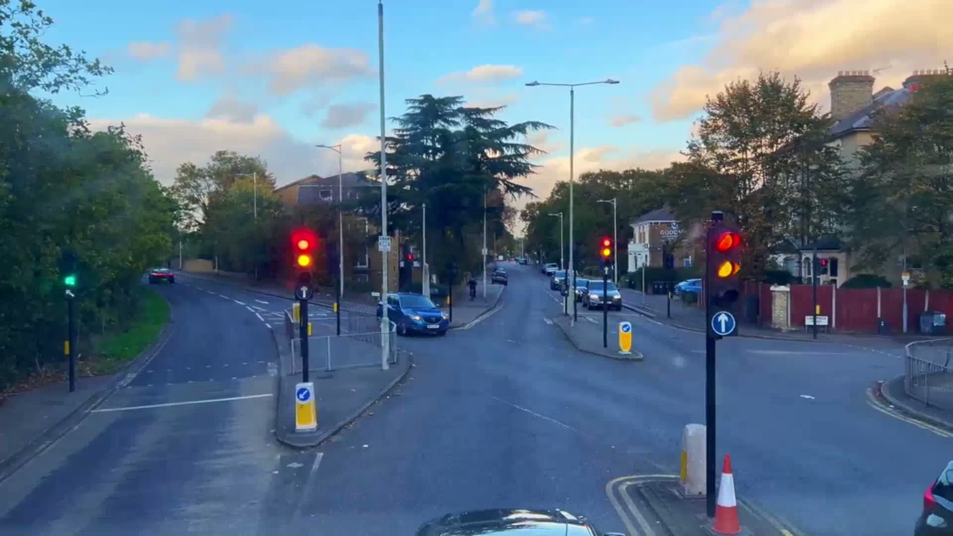 A mostly clear, late afternoon scene shows a residential street intersection with several cars stopped at traffic signals under a partly cloudy sky.