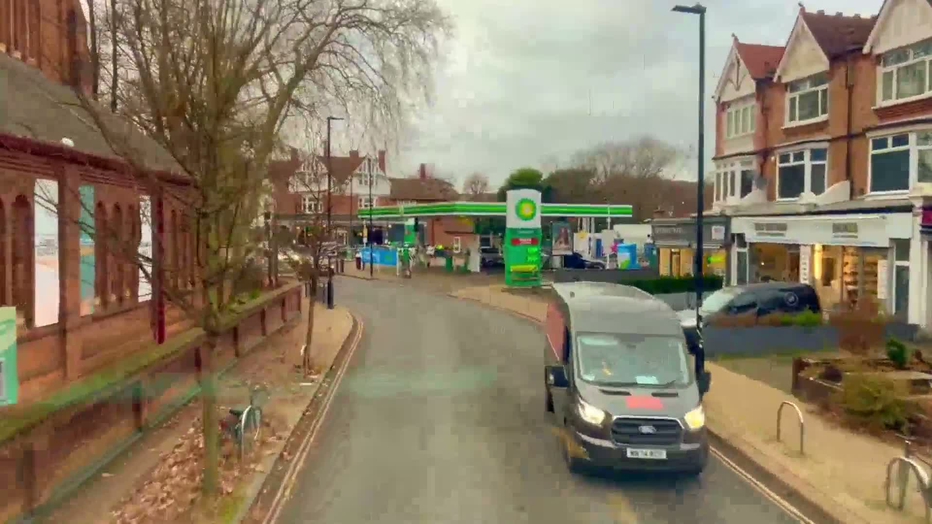 A dark-grey van drives down a street past brick buildings and a gas station on an overcast day.