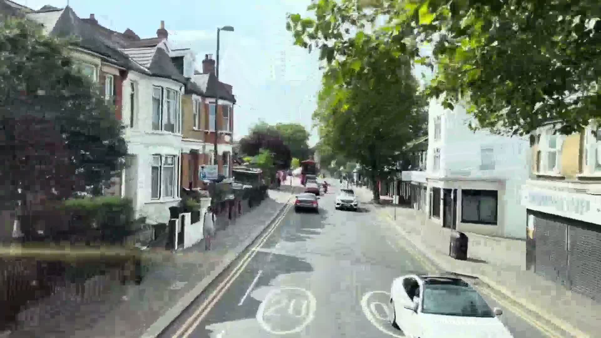 A street lined with houses and shops is seen from a moving vehicle on a sunny day.