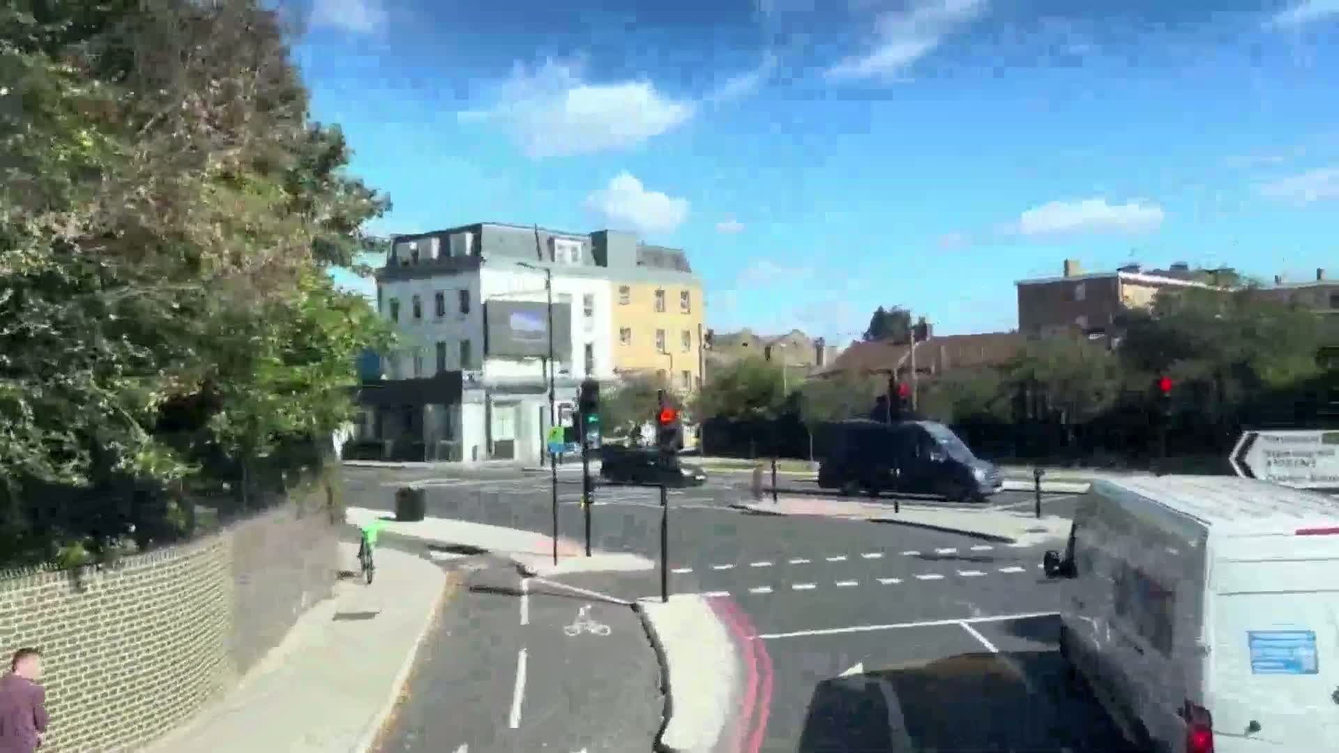 A sunny day shows a busy intersection with several vehicles, pedestrians, and cyclists, surrounded by buildings and lush green trees.