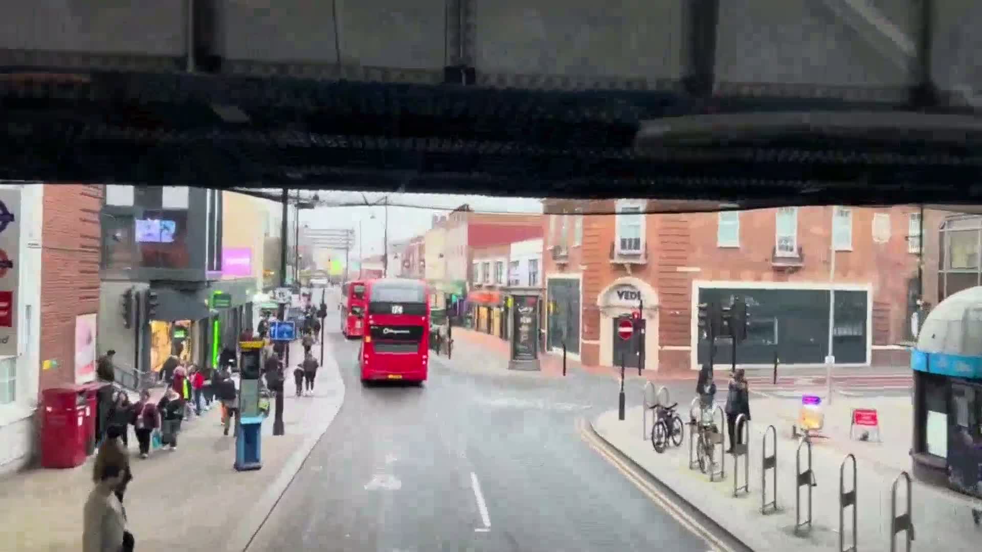A view from inside a bus shows a rainy street scene with pedestrians and two double-decker buses, one in the distance and one closer to the camera.