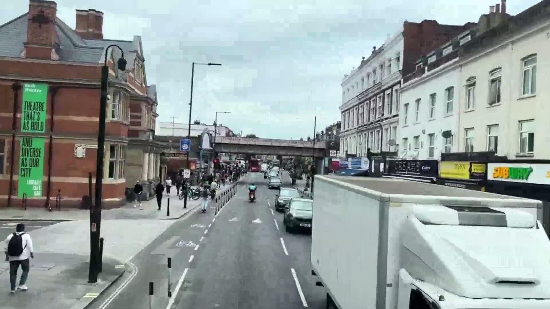 A city street scene on an overcast day shows pedestrians and vehicles, including a large white truck, moving along a road lined with buildings.