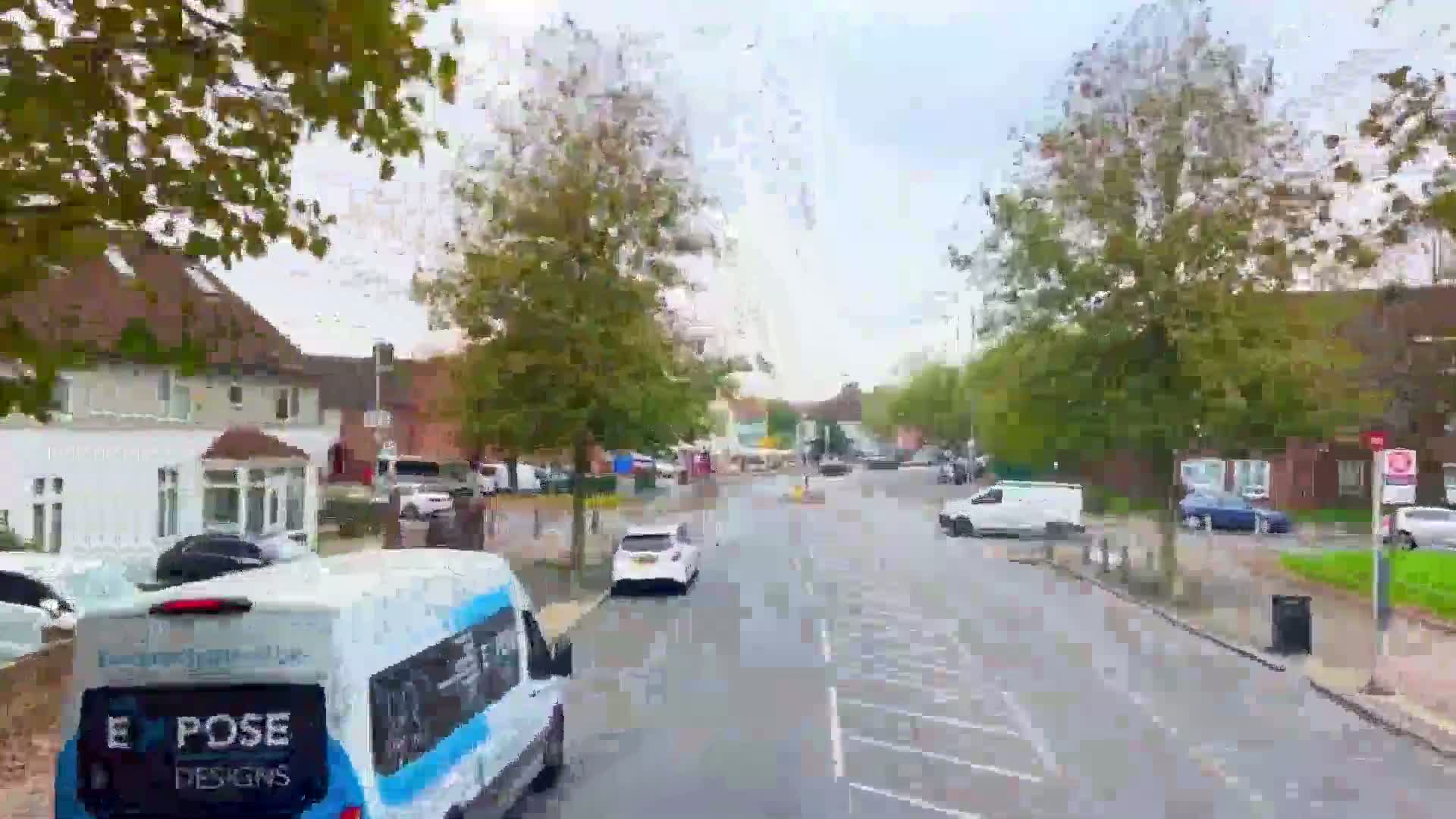 A wet street lined with houses and trees, showing several parked and driving cars on a cloudy day.