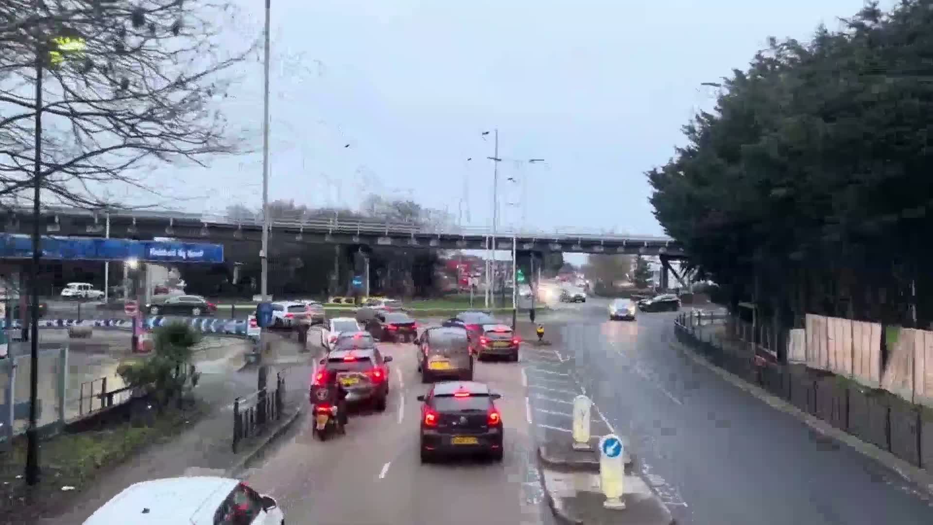 A wet, overcast day shows traffic driving on a multi-lane road that passes under a bridge and through a roundabout.