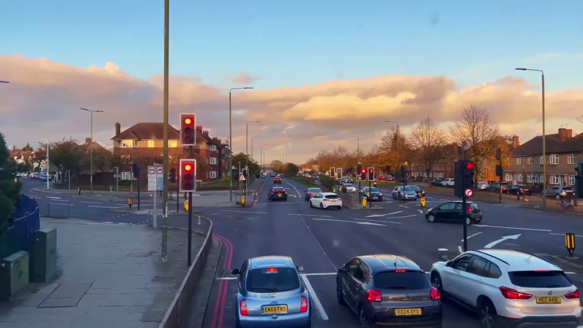 Under a partly cloudy, sunset sky, cars are stopped at a multi-lane intersection in a residential area with buildings lining both sides of the road.