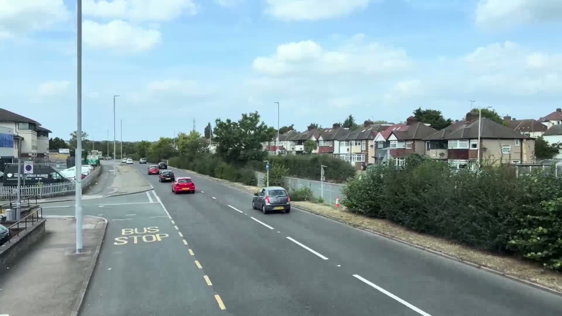 A mostly sunny day reveals a road with several cars driving past a row of houses and a hedgerow.