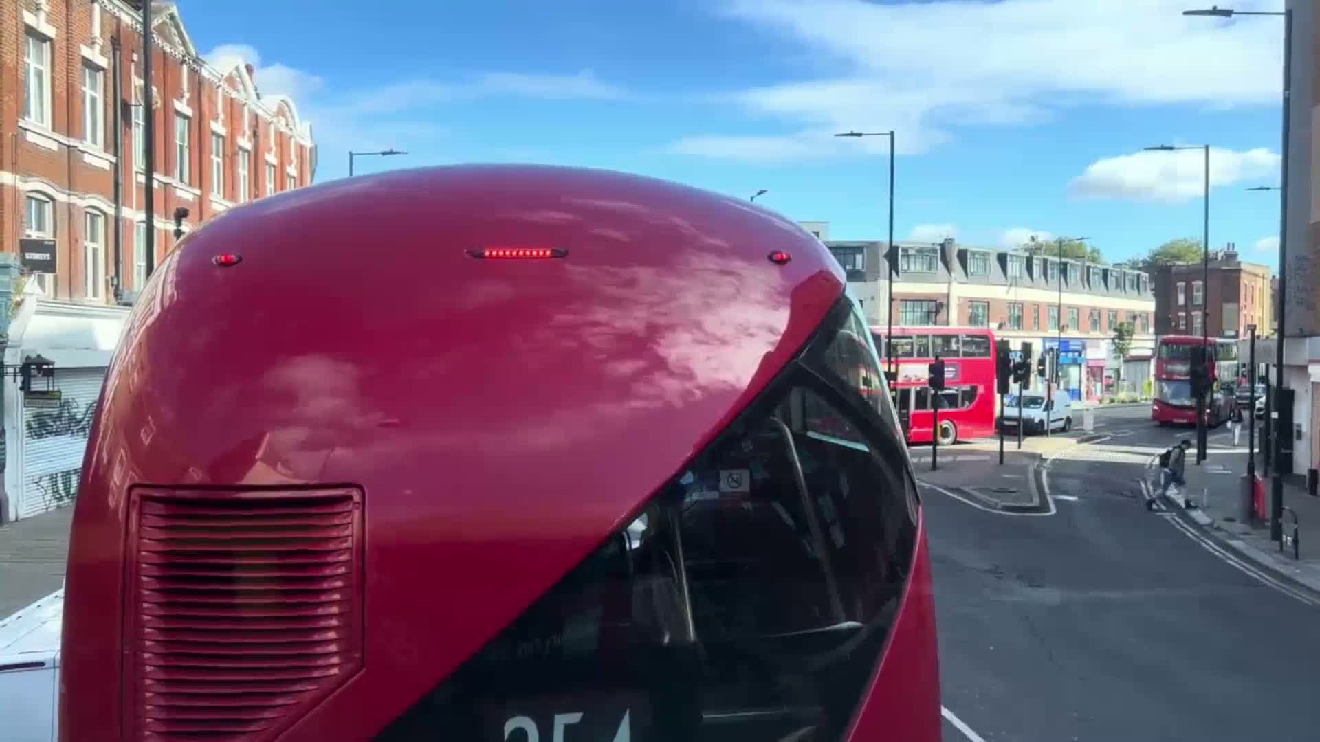 The rear of a red double-decker bus is shown on a sunny day, with other buses and buildings in the background.
