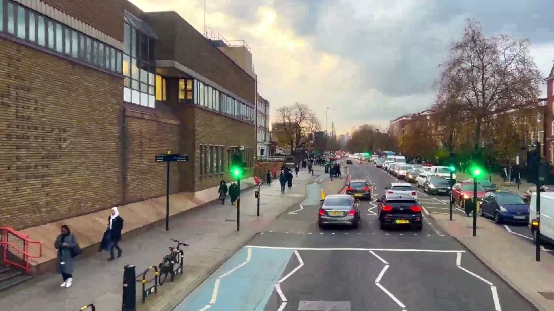 A city street scene with overcast skies shows pedestrians walking on sidewalks and cars driving on a road that passes brick buildings and bare trees.