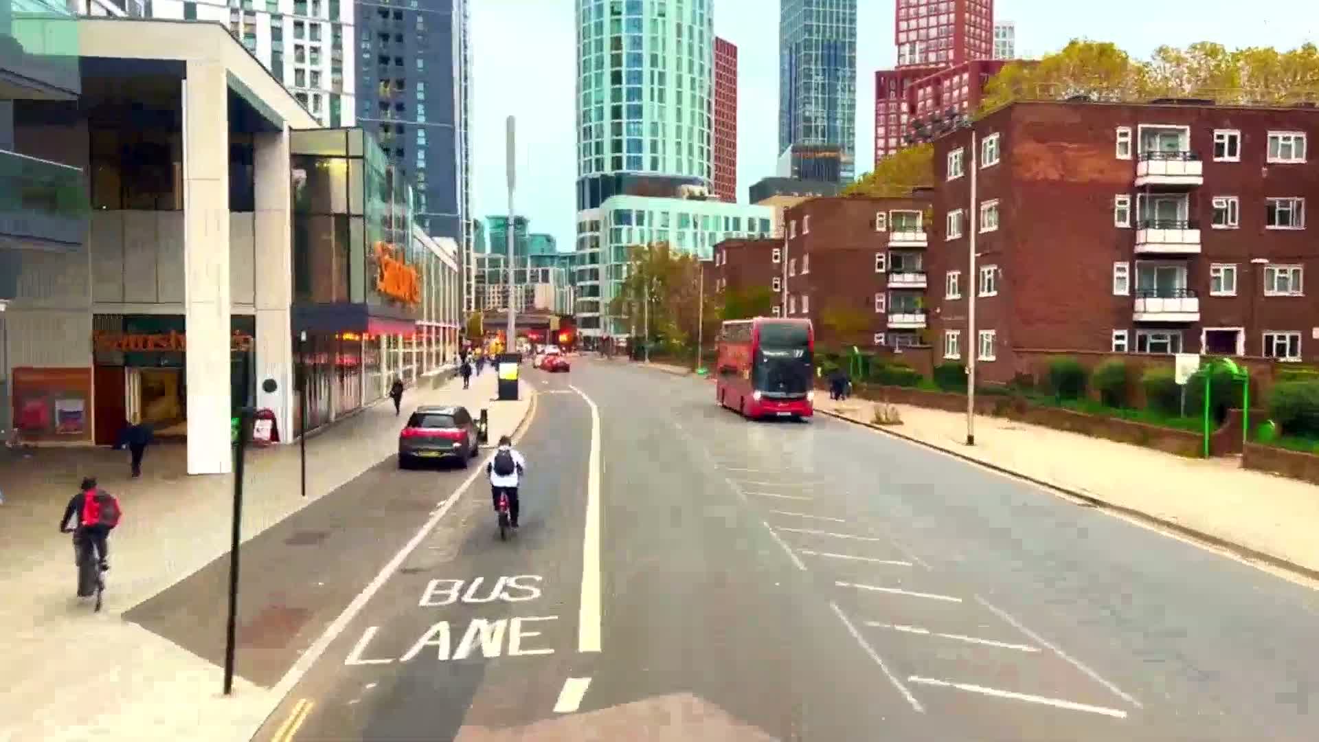 A cyclist rides along a city street with a double-decker bus and several cars, under a partly cloudy sky, past modern and older buildings.