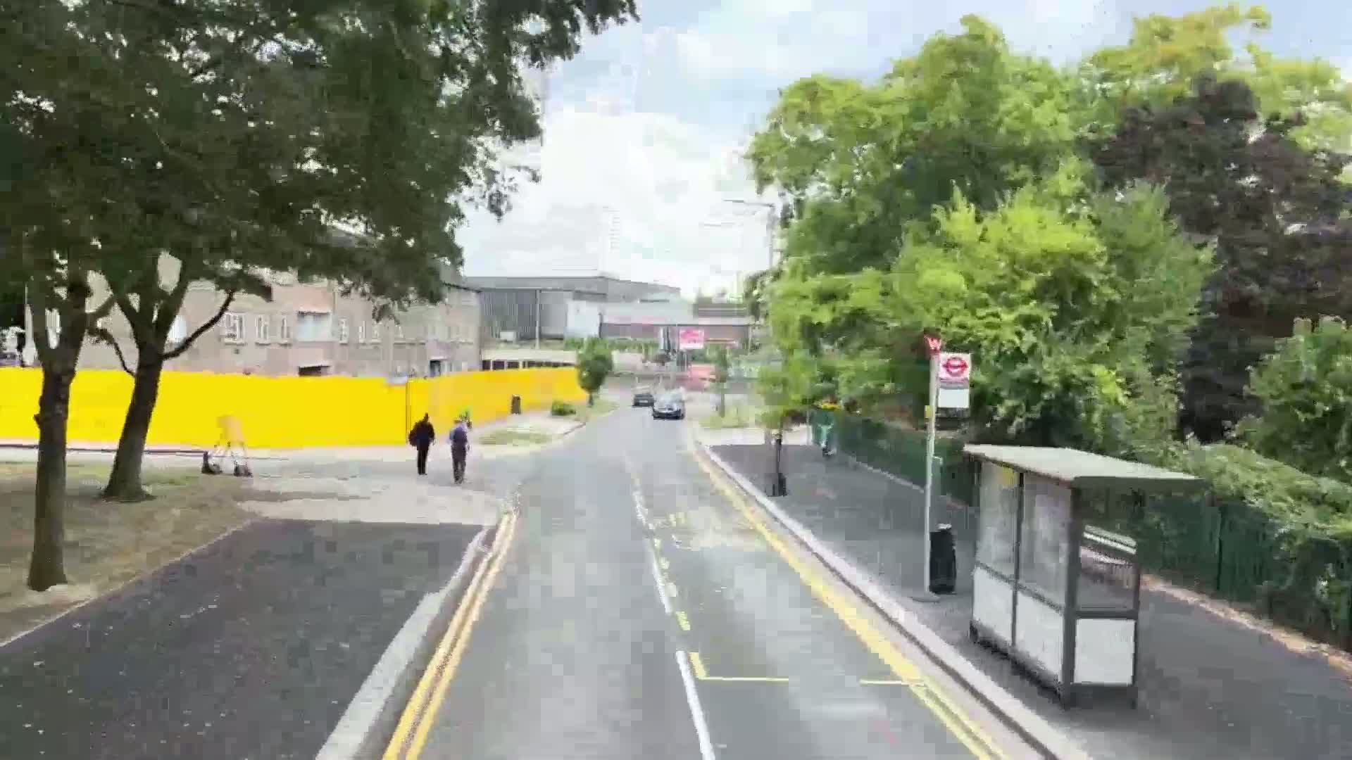 A street scene on a partly cloudy day shows a two-lane road with a bus shelter, pedestrians, and a few cars; a yellow construction fence is visible in the background.