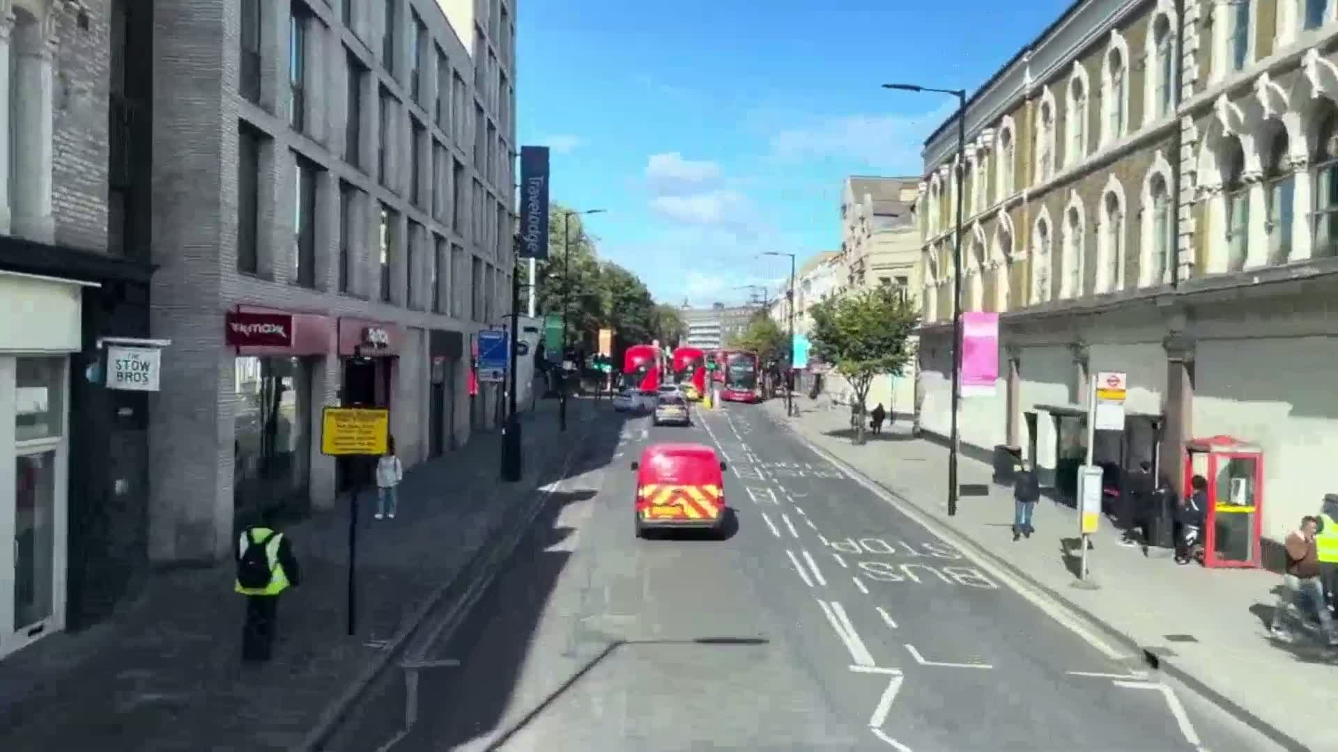 A sunny city street scene shows several vehicles including buses and vans traveling along a road lined with buildings and pedestrians on a sunny day.