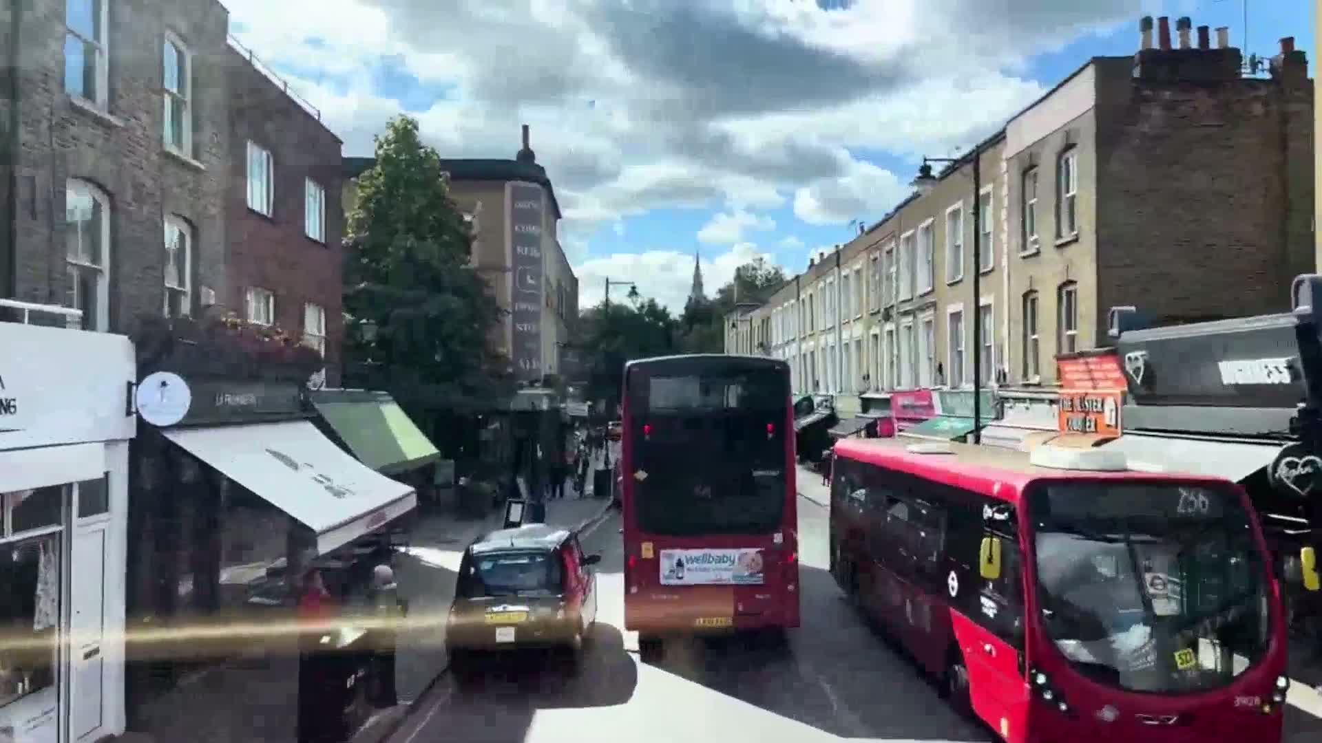 A sunny day on a city street shows several buses and cars driving past brick buildings with shops on the ground floor.