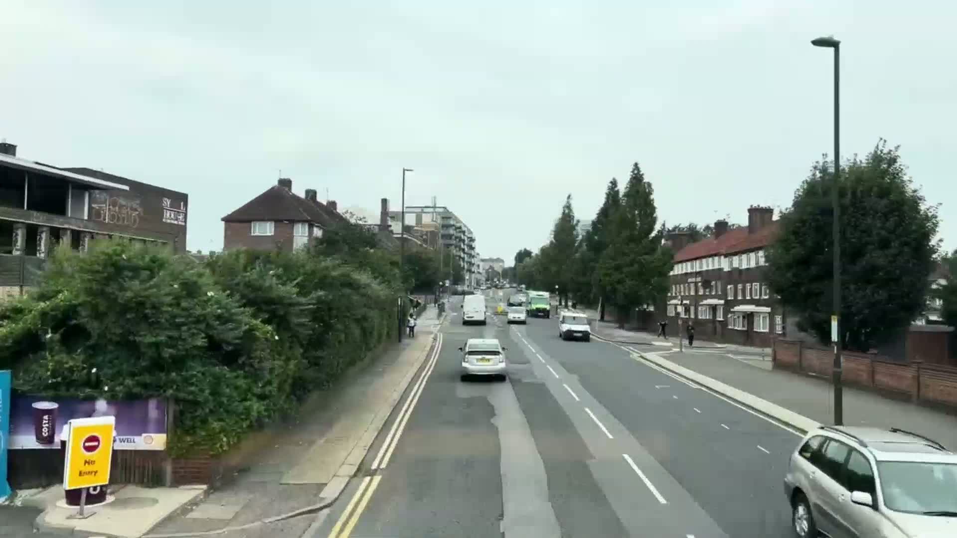 A street scene on an overcast day shows several cars driving on a multi-lane road lined with residential buildings and lush green hedges.