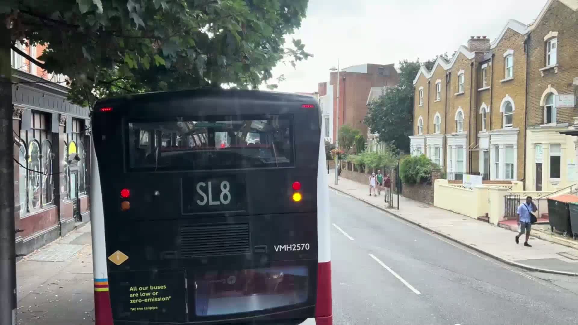 A view from the back of a bus driving down a residential street on a cloudy day, with pedestrians walking on the sidewalk.