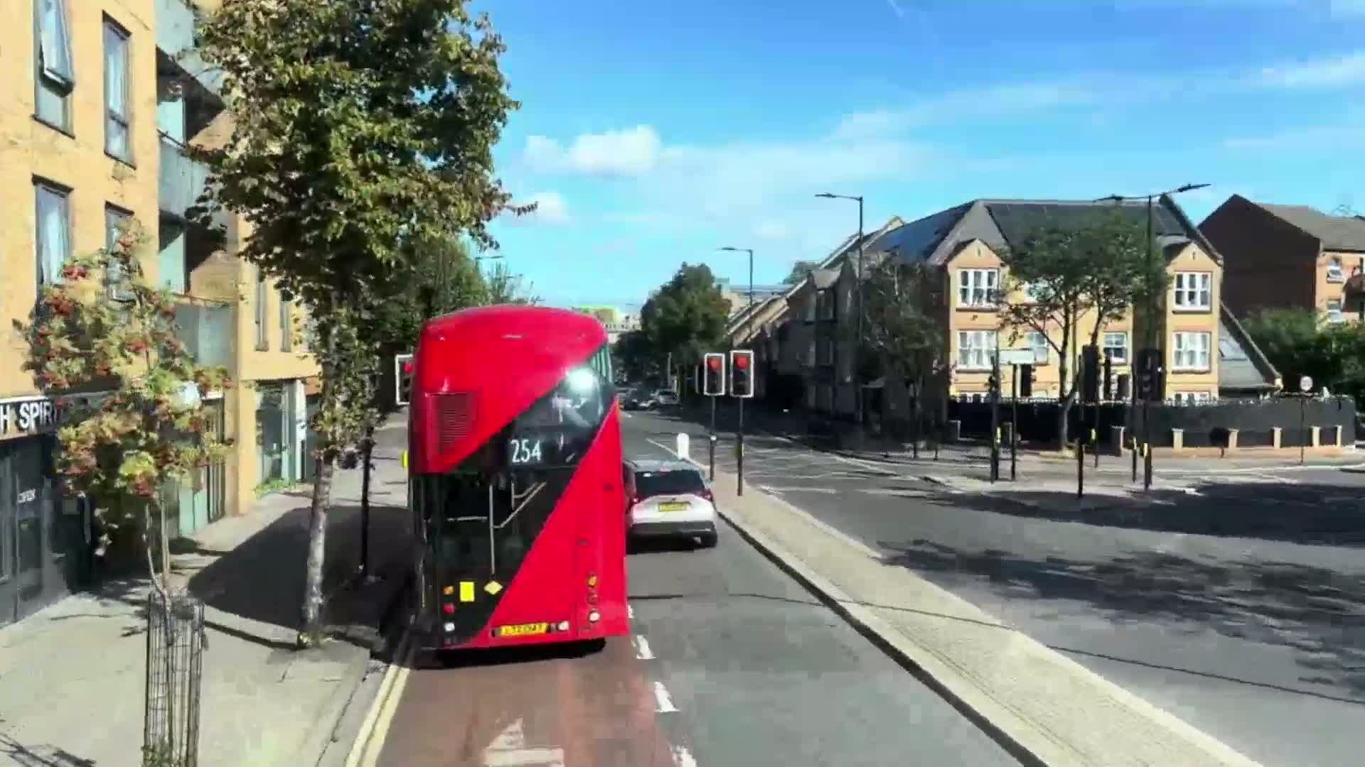 A red double-decker bus is stopped at a traffic light on a sunny day in a residential area, with cars in front and buildings lining the street.