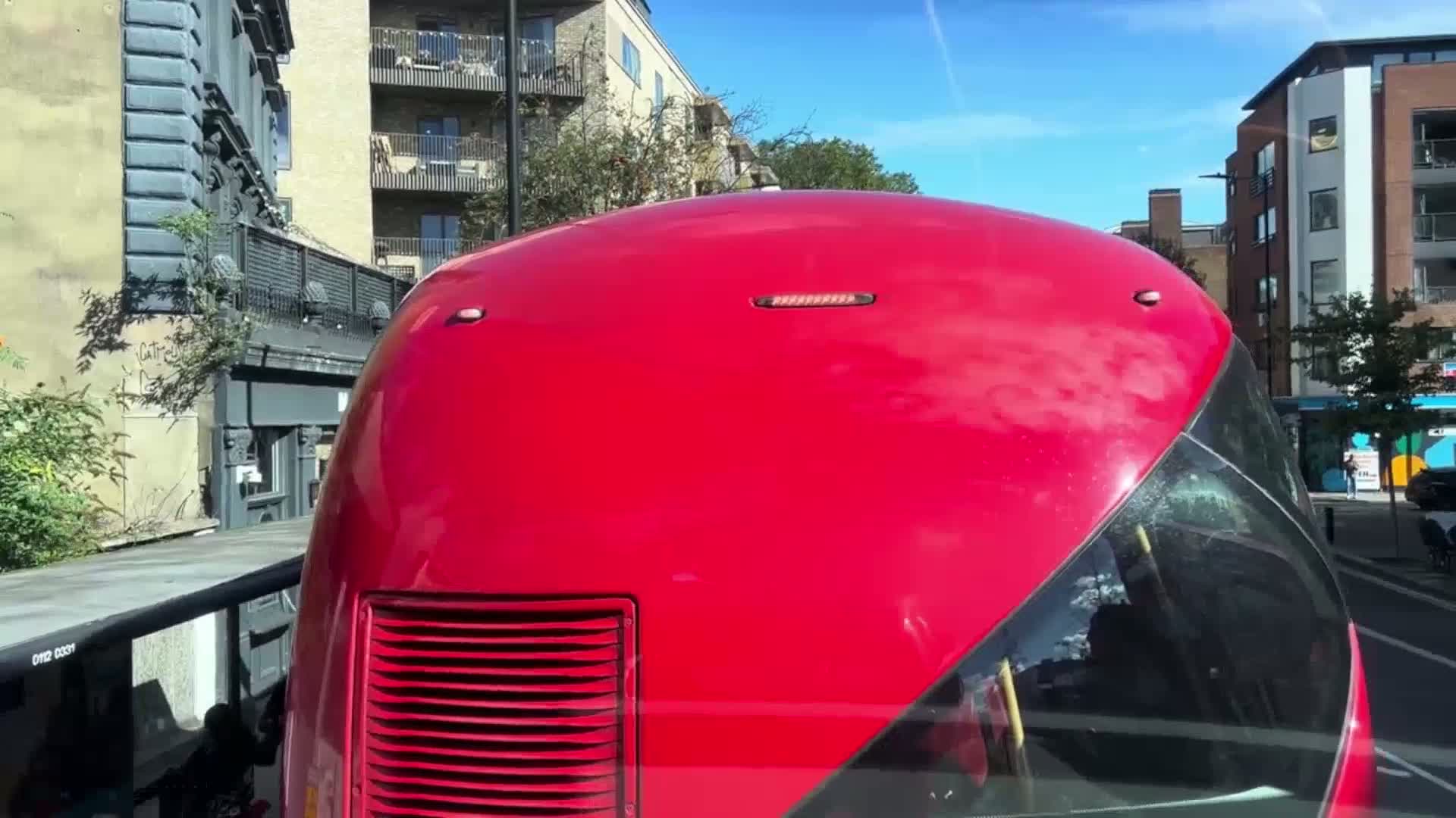 The rear of a bright red bus is visible, traveling on a sunny day past buildings.