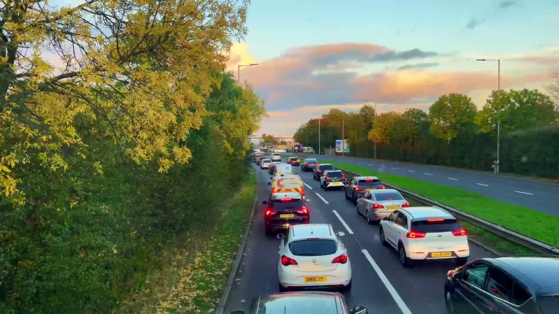 A traffic jam on a multi-lane road at sunset, under a partly cloudy sky, with autumnal trees lining the roadside.