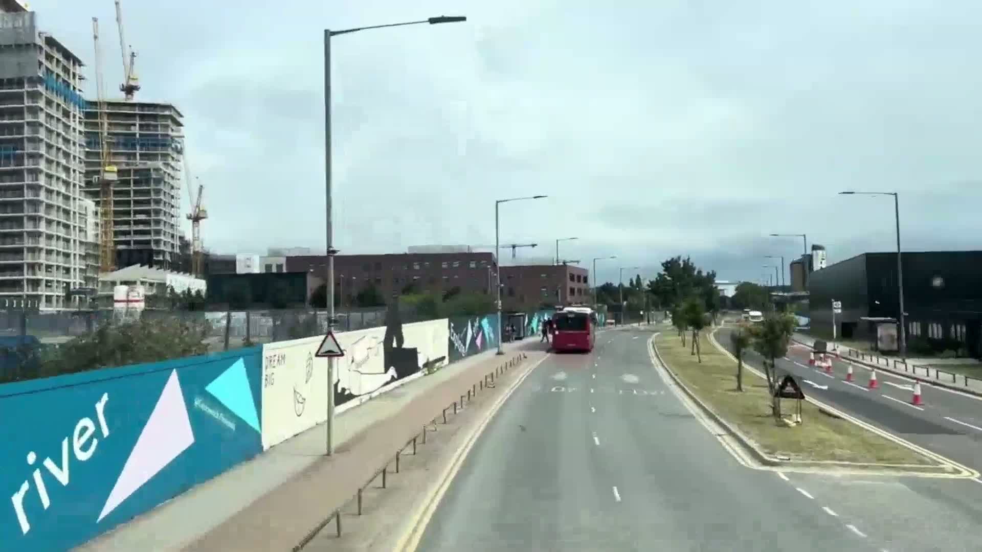 A red bus drives down a multi-lane road on an overcast day, passing by a construction site with tall buildings and a colorful mural-covered wall.