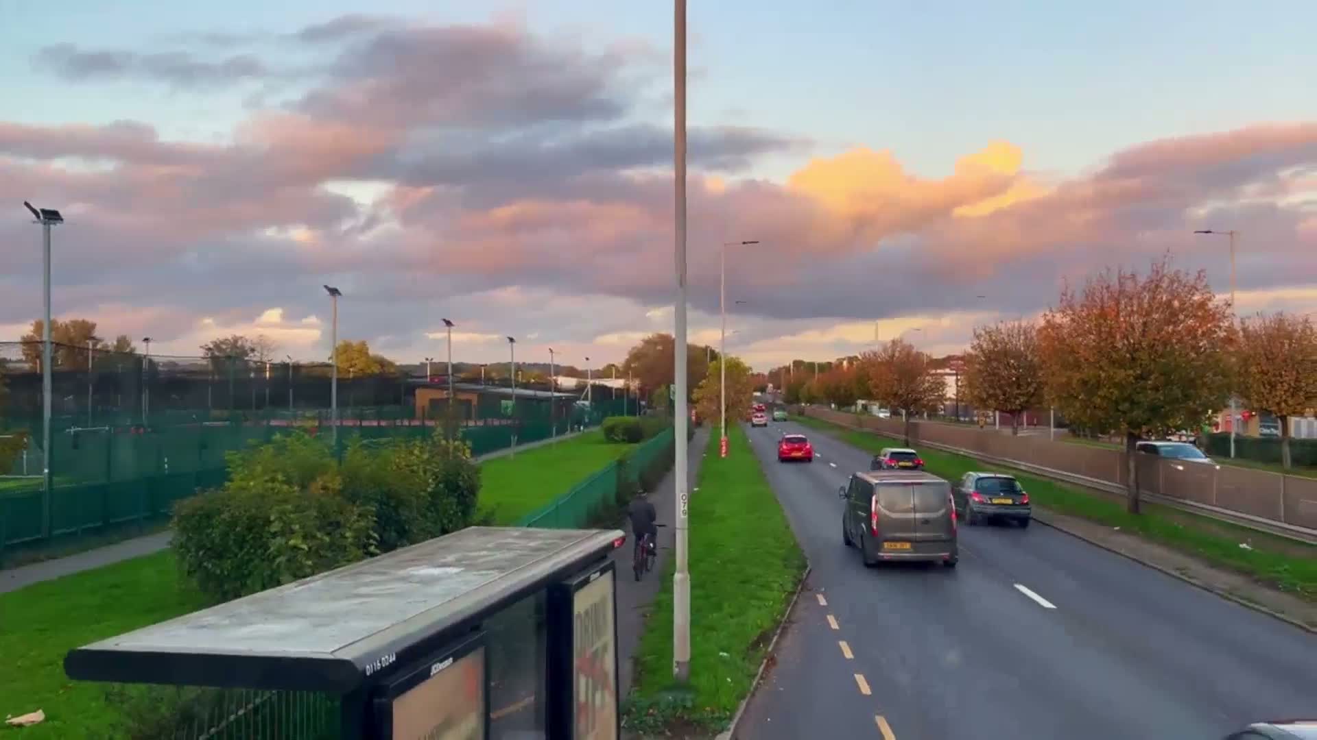 A multi-lane road with several cars driving under a partly cloudy sky displaying pink and orange hues at sunset, alongside a grassy area with tennis courts and trees.