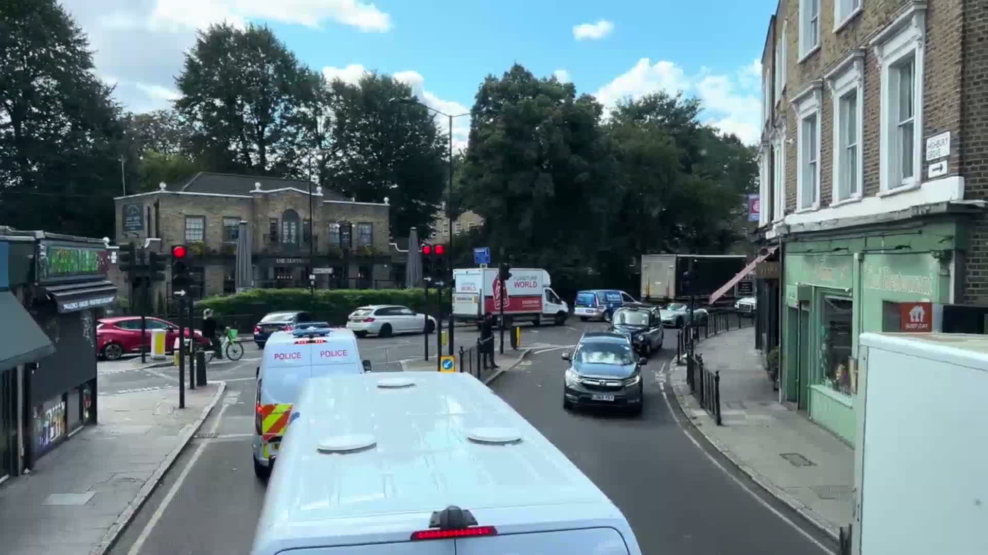 A sunny day shows several cars and a police van at a crossroads near some shops and a pub, with trees lining the street.