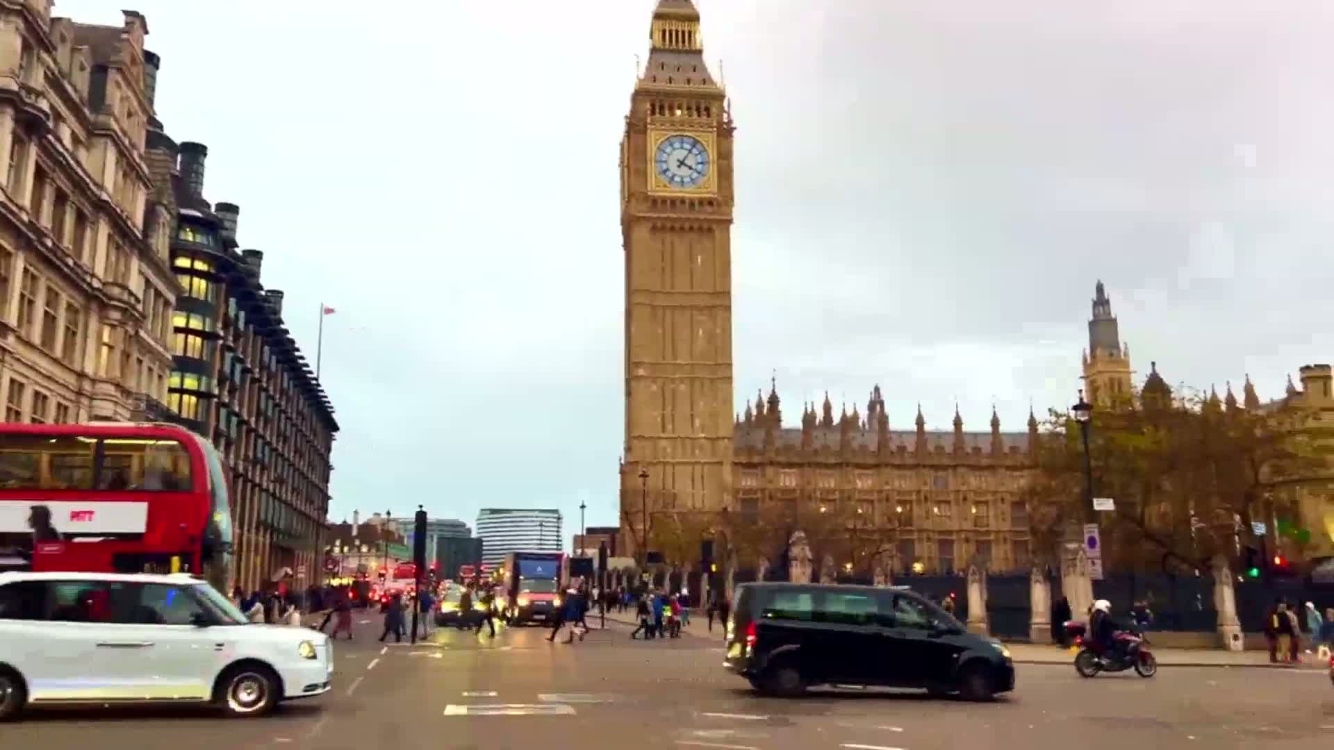 A city street scene shows Big Ben and the Houses of Parliament in the background, with pedestrians and vehicles including a double-decker bus and a white taxi-like car in the foreground, under an overcast sky.