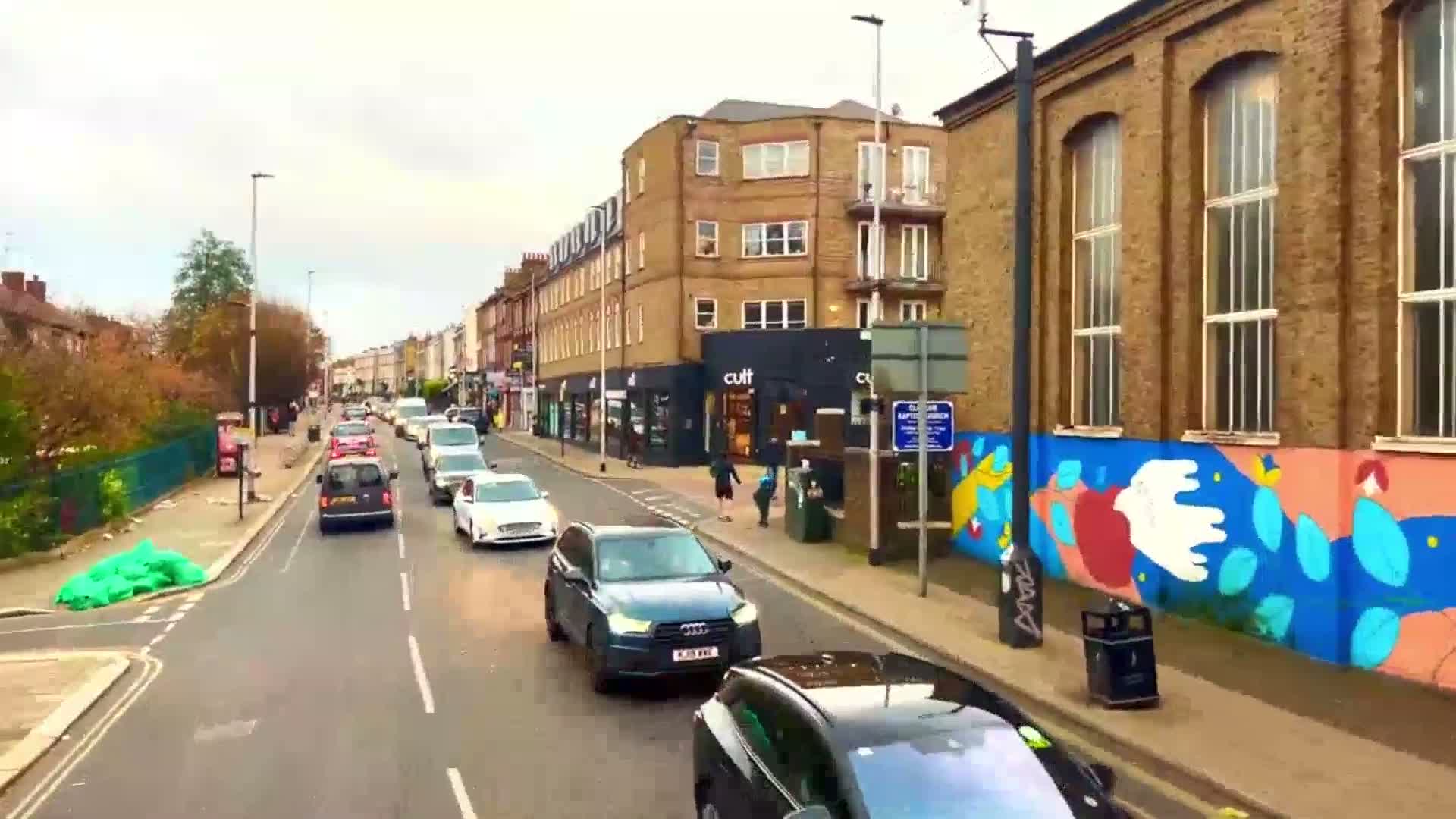 A street scene on an overcast day shows several cars driving past multi-story buildings, one of which has a colorful mural on its side.