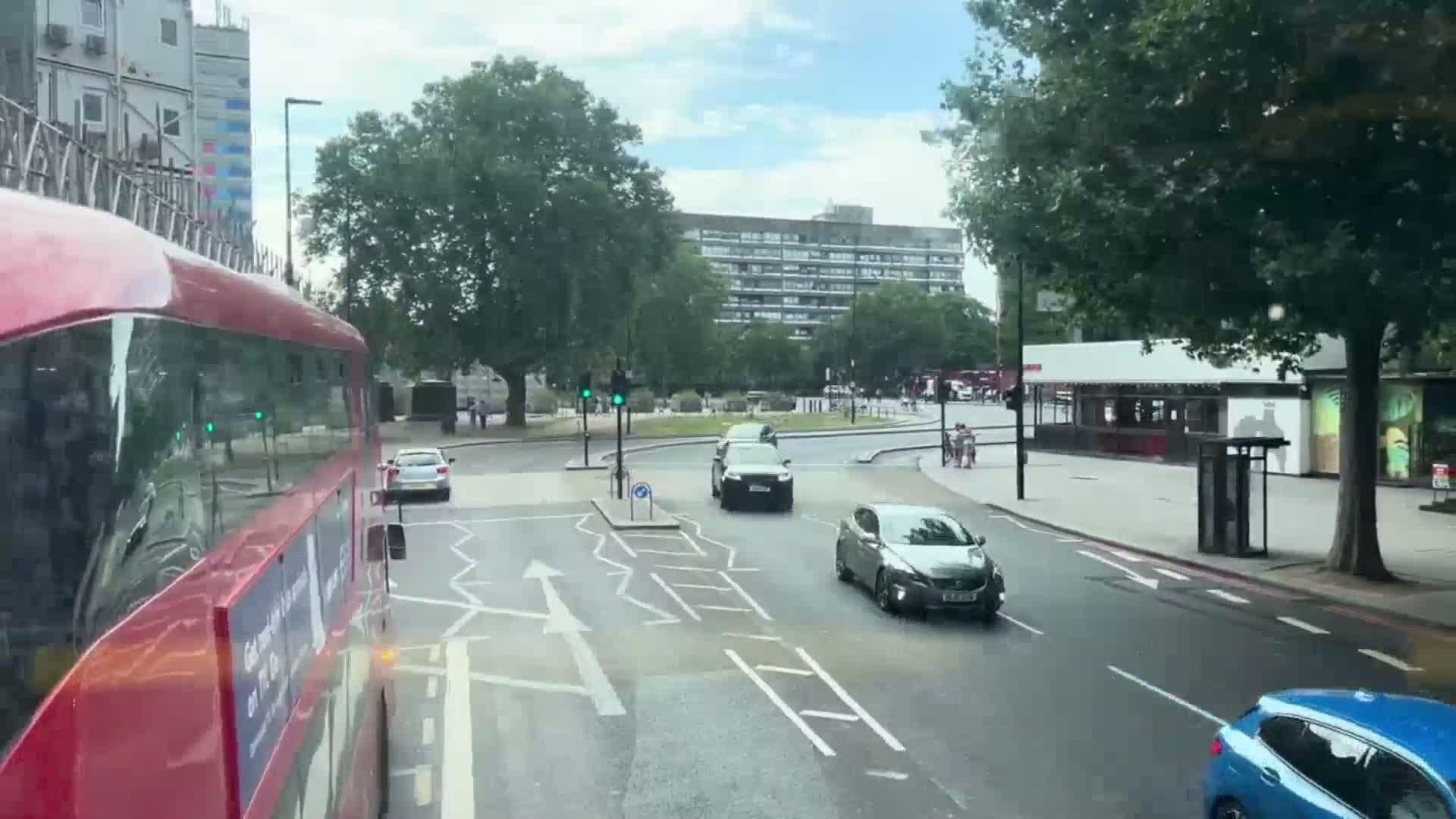 A view from a double-decker bus shows a city street scene on a partly sunny day, with several cars at a traffic intersection and pedestrians walking on the sidewalk.