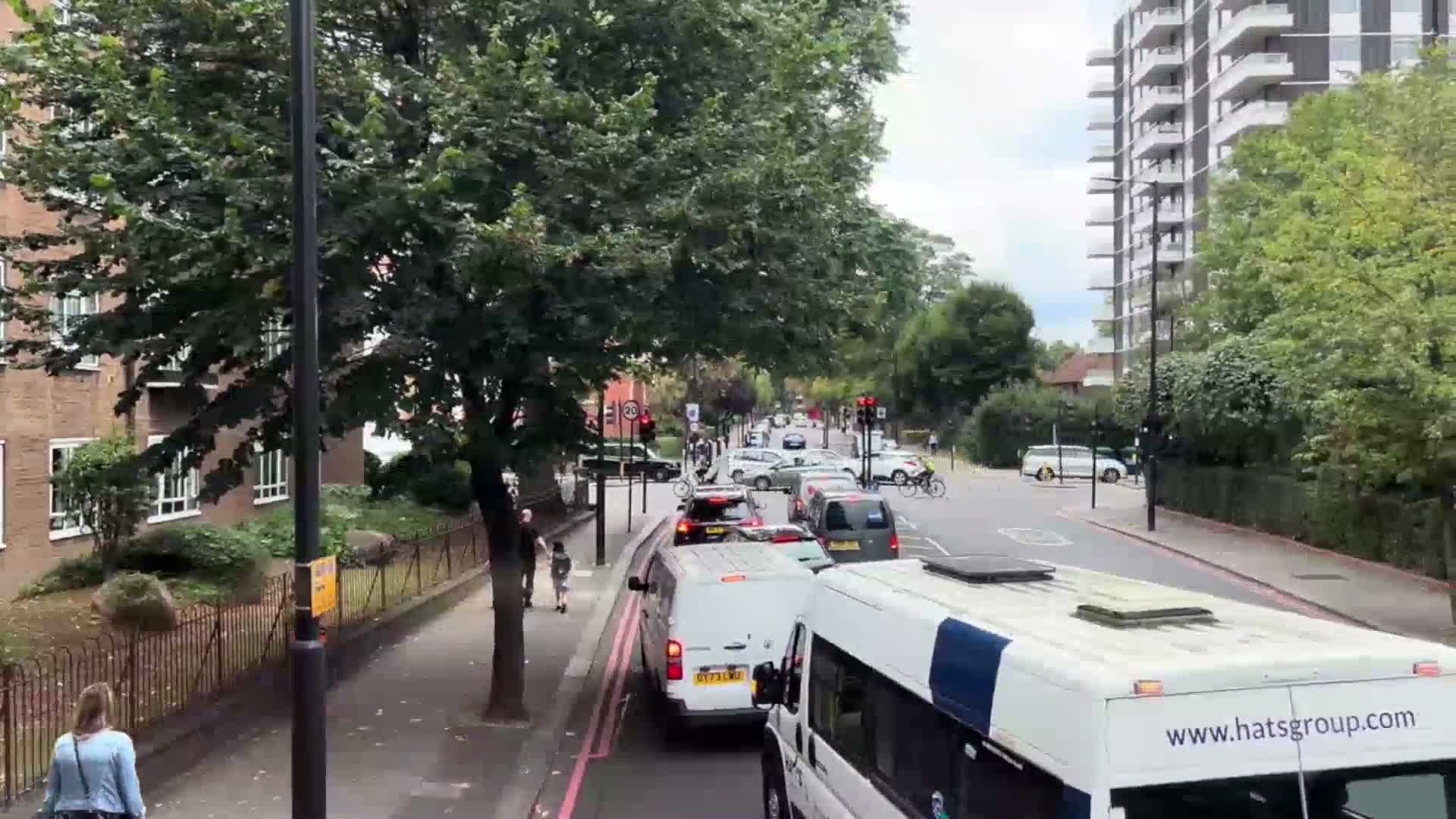 A partly cloudy day reveals a city street scene with moderate traffic congestion, including cars, vans, and cyclists, lined with buildings and trees.