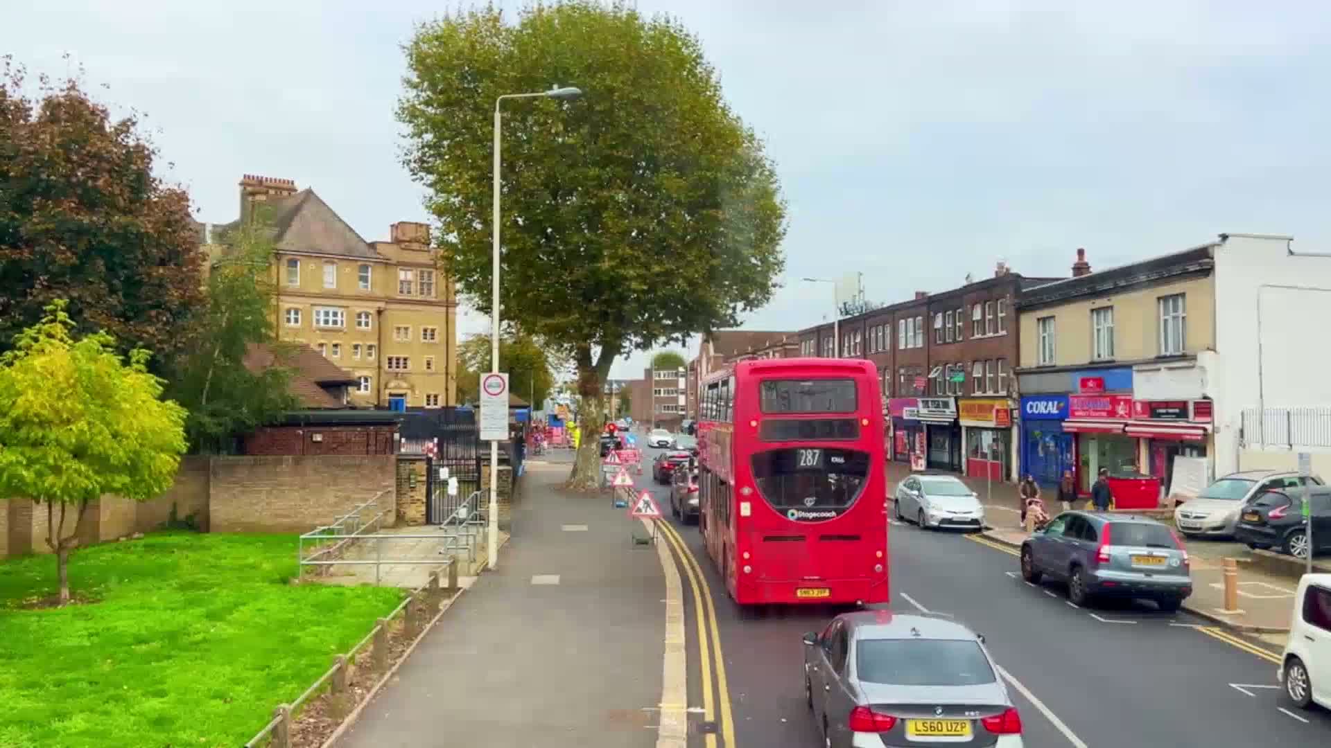A red double-decker bus drives down a street lined with multi-story buildings and shops under an overcast sky.