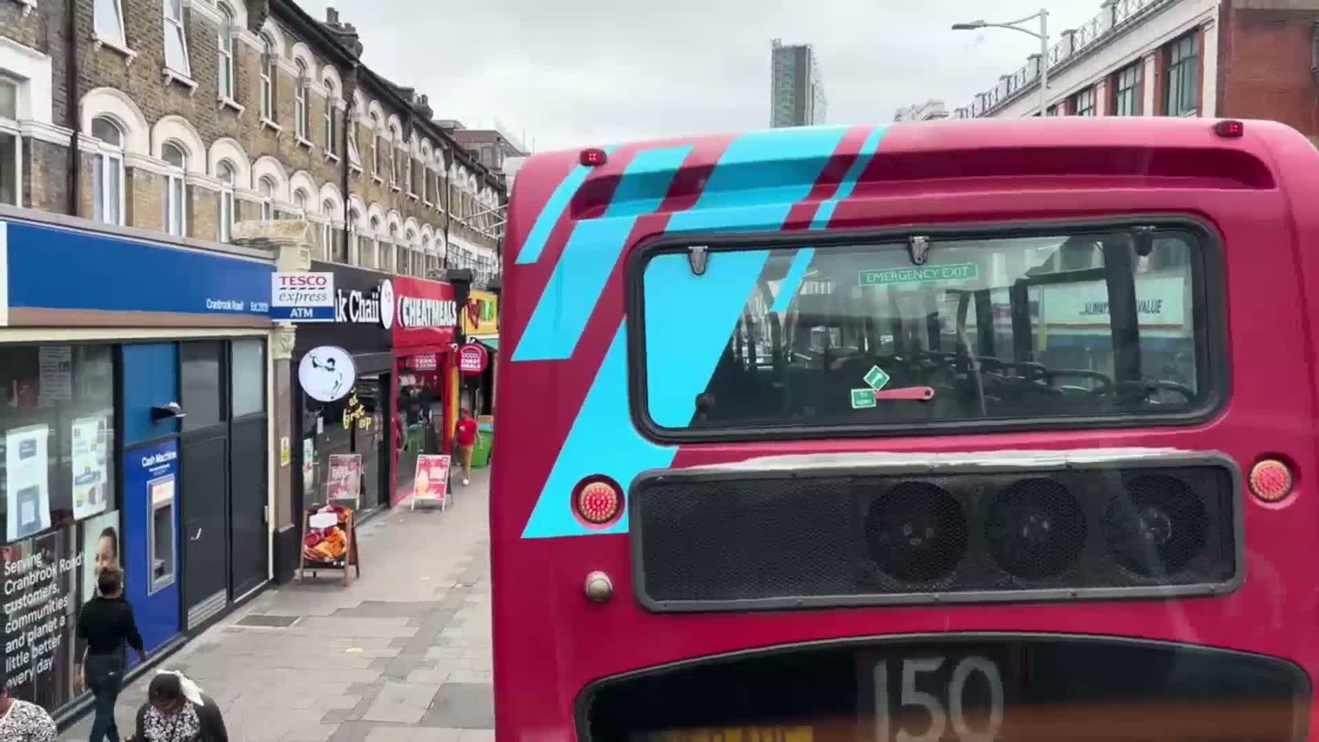 The rear of a red double-decker bus with teal accents is visible, driving down a street lined with shops on an overcast day.