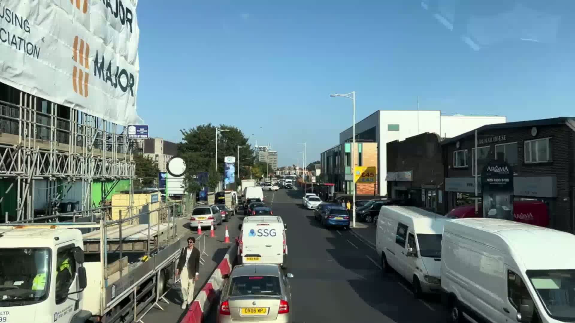 A sunny day reveals a city street scene with moderate traffic, construction underway on a building to the left, and various shops and buildings lining the road.