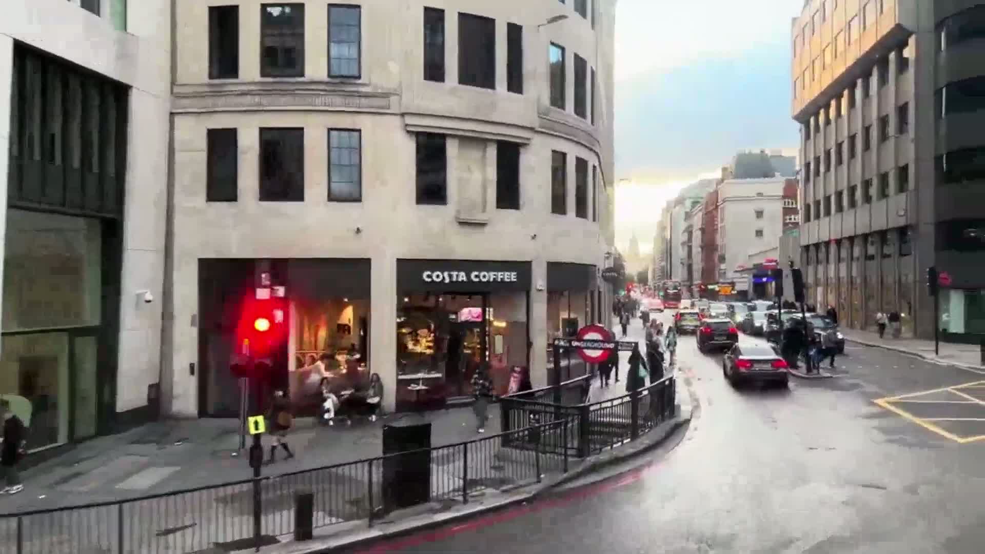 A city street scene shows pedestrians and cars on a wet road, with a Costa Coffee shop and a red traffic light visible in the foreground, under an overcast sky.
