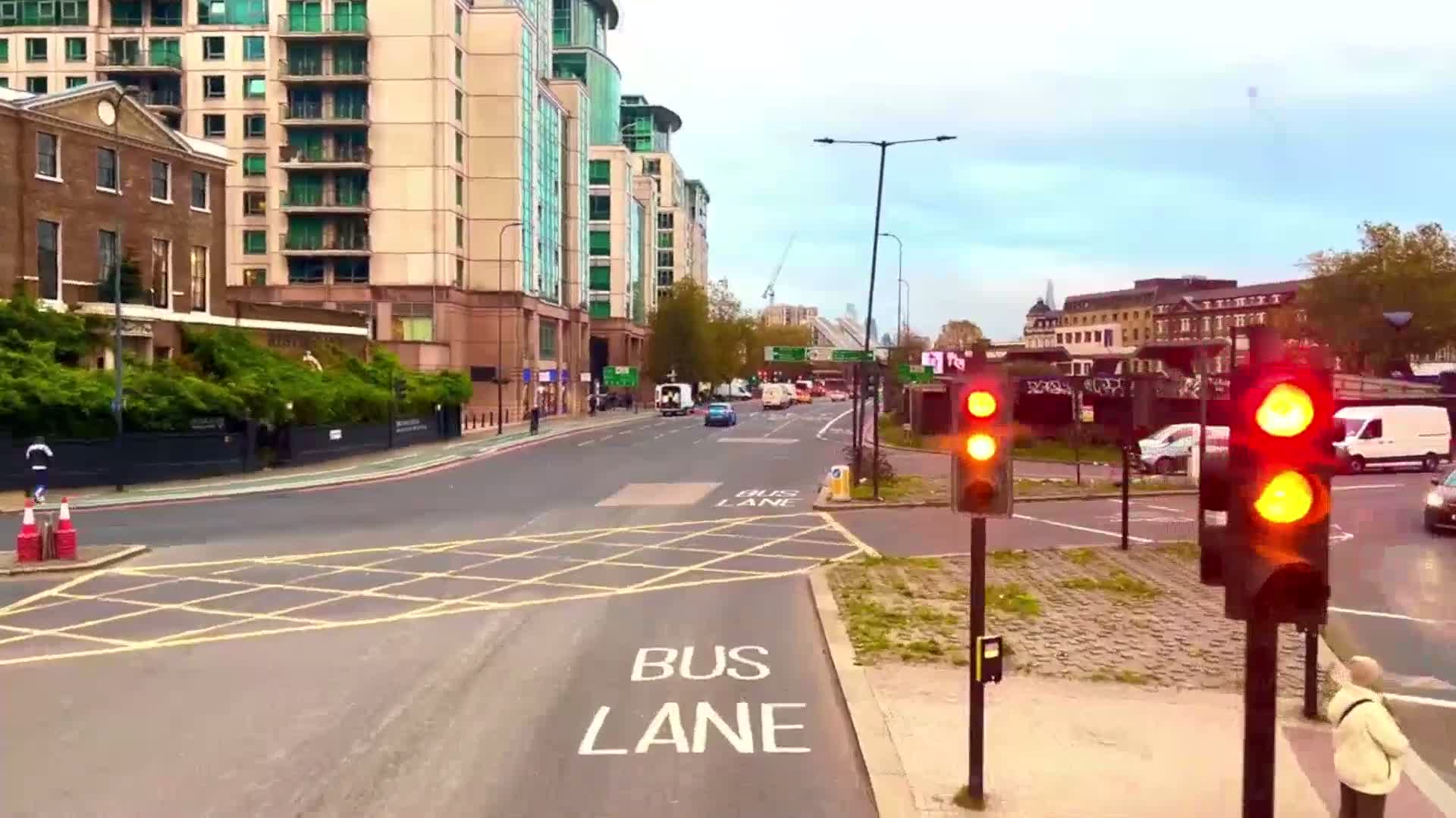 A city street scene under a partly cloudy sky shows a bus lane intersection with traffic lights displaying red, several vehicles, and buildings lining the street.