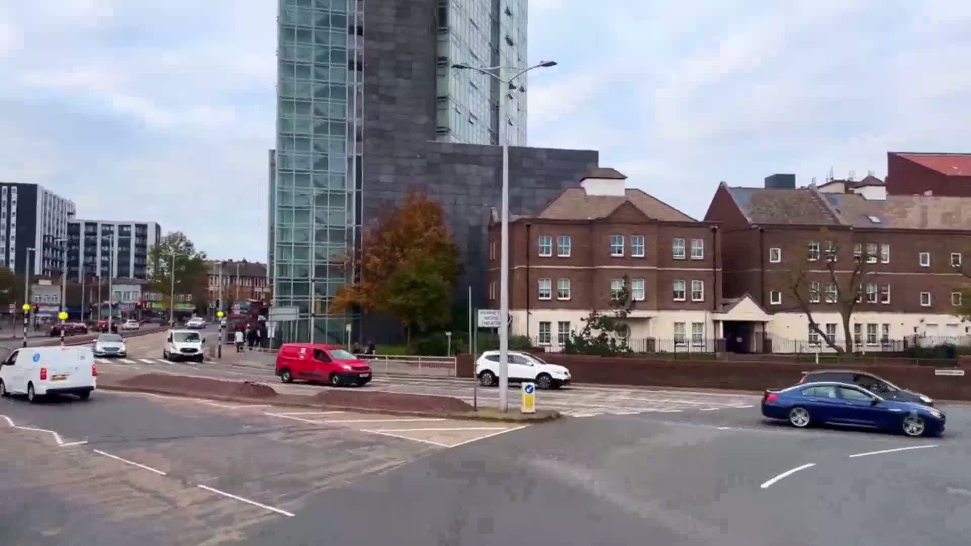 Several vehicles are driving around a roundabout in a city with overcast skies, near a tall glass building and several brick buildings.