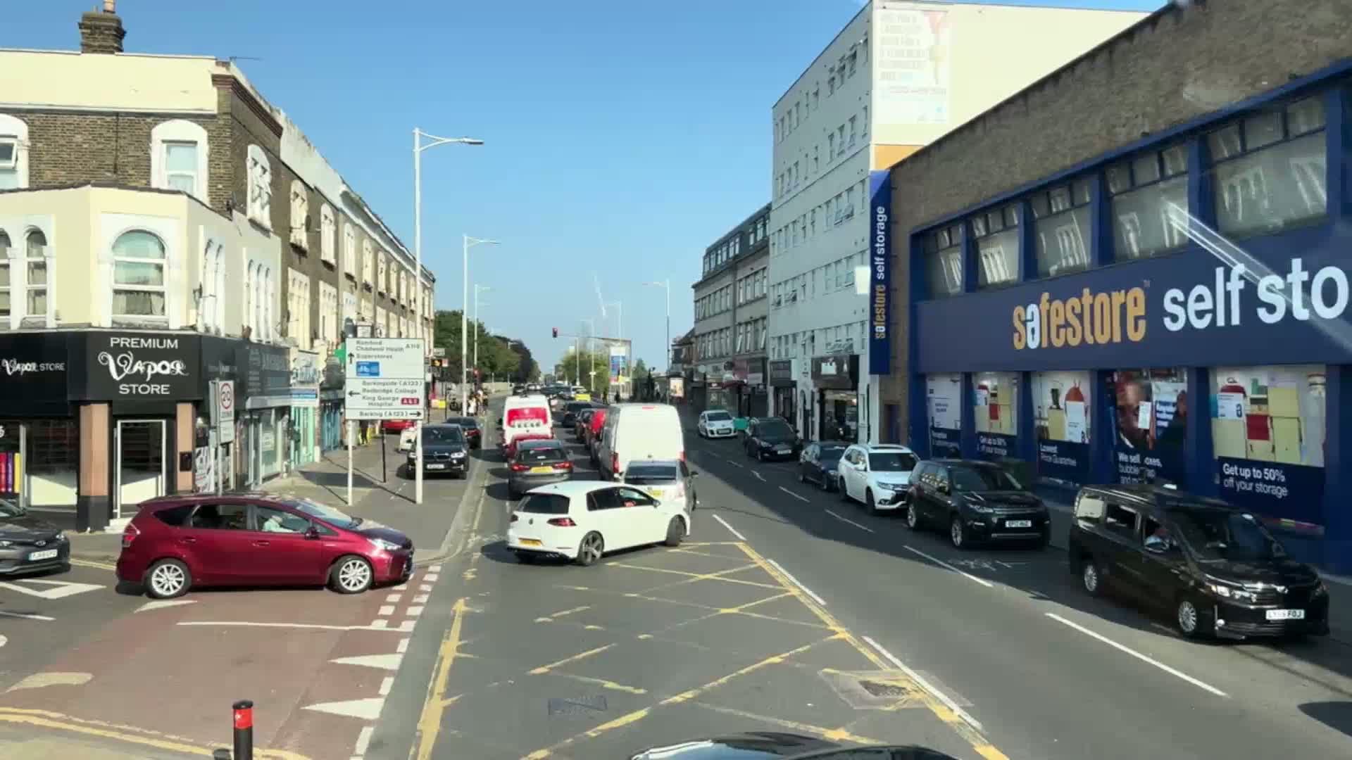 A sunny day reveals a busy, multi-lane road with moderate traffic, lined with shops and buildings on either side.