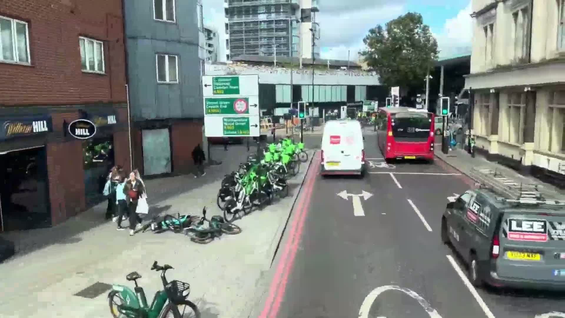 A city street scene on a sunny day shows a row of green bicycles parked at the curb, several pedestrians walking, a white van and a red bus in traffic, and a gray van partially visible in the foreground.