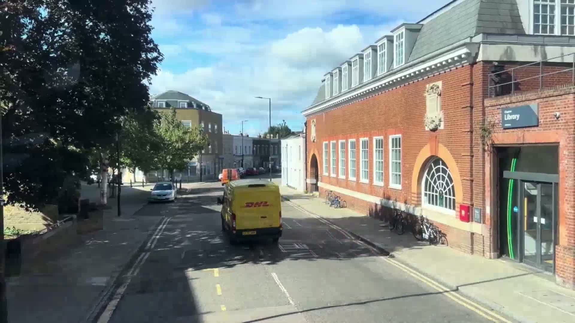 A yellow delivery van drives down a sunny street lined with brick buildings and trees, one of which is a library.
