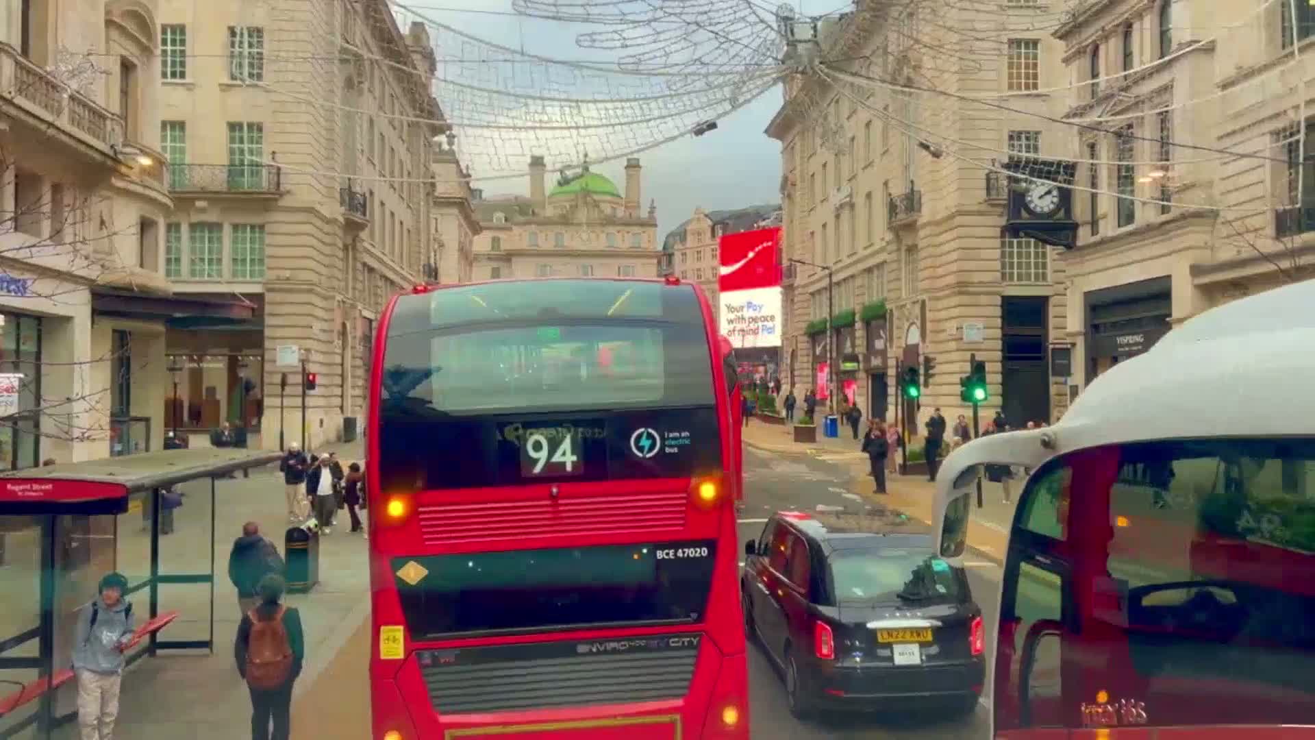 A red double-decker bus travels down a city street lined with buildings adorned with holiday decorations, under an overcast sky.
