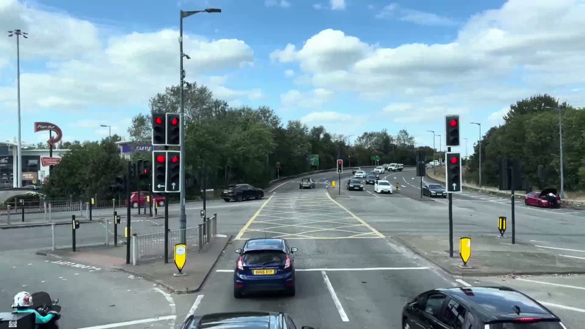 A busy road intersection with traffic lights showing red, several cars stopped and moving, under a partly cloudy sky.