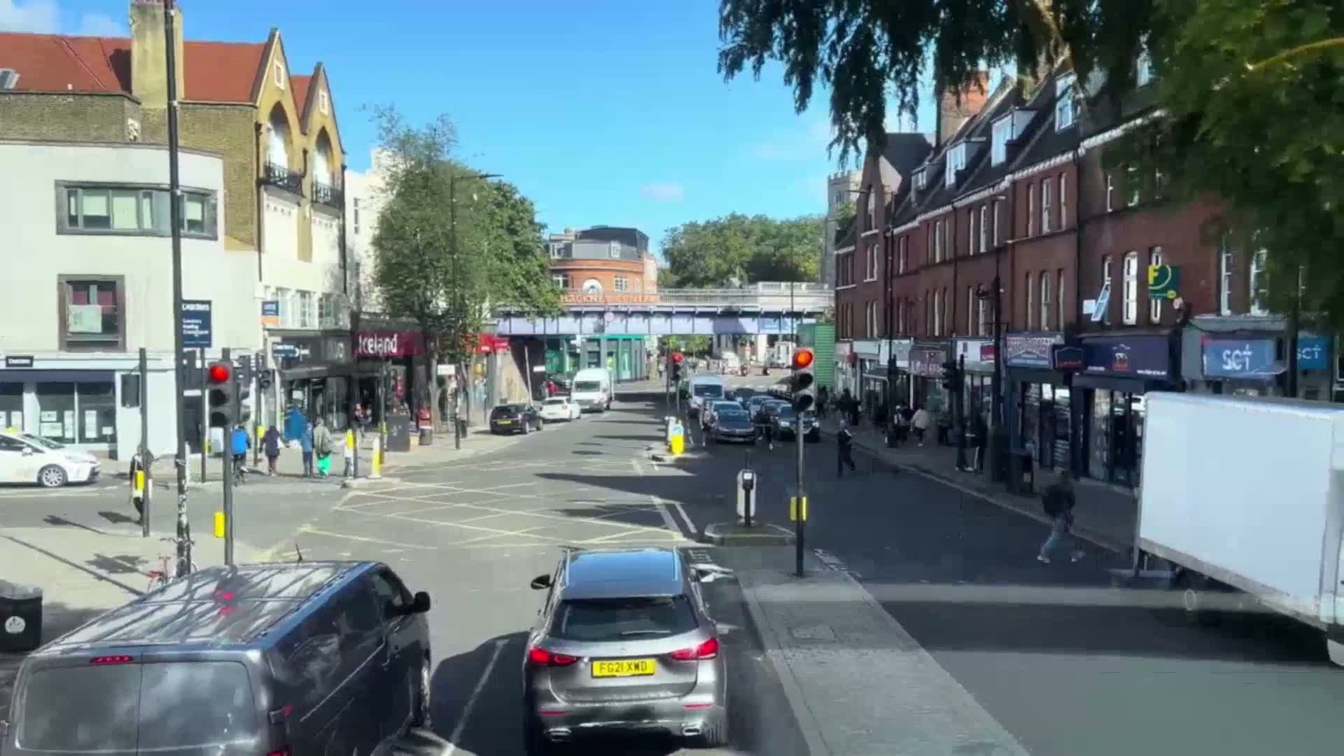A sunny street scene shows several cars and pedestrians at a crossroads, with multi-story buildings lining both sides of the street under a mostly clear sky.