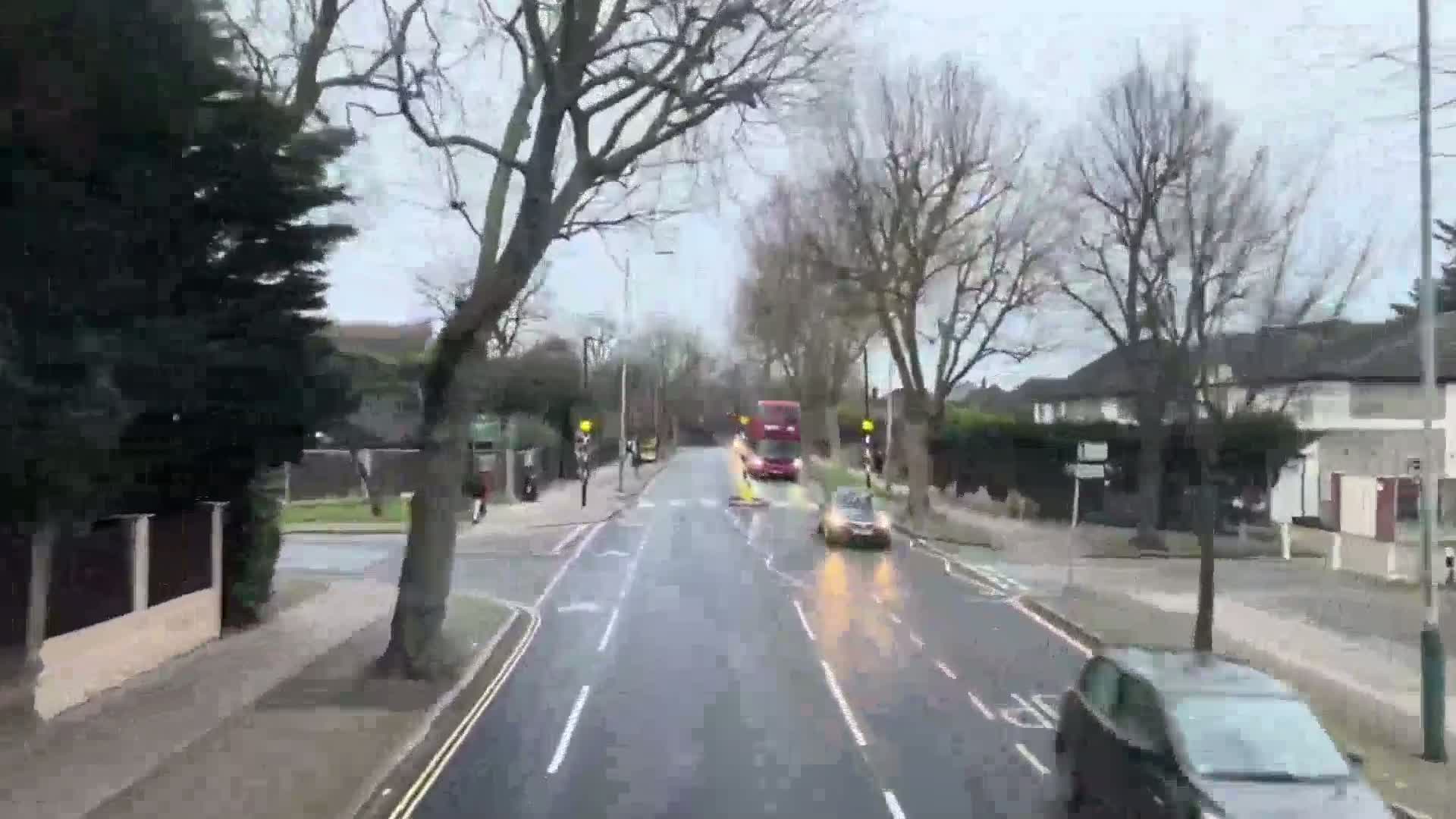 A wet, overcast day reveals a residential street with bare trees lining a road where a double-decker bus and several cars are driving.
