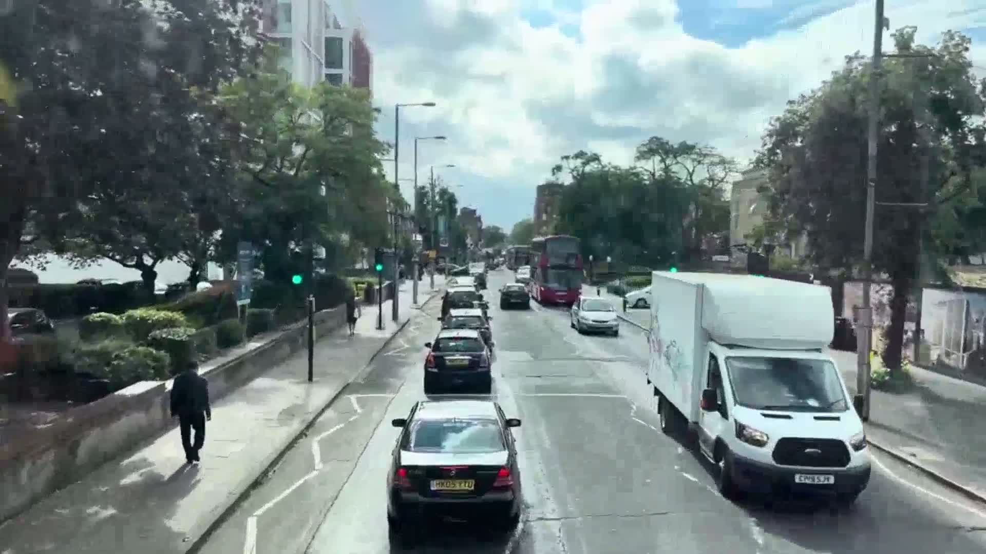 A partly cloudy day shows a line of cars and a van driving down a tree-lined city street, with a pedestrian walking on the sidewalk.