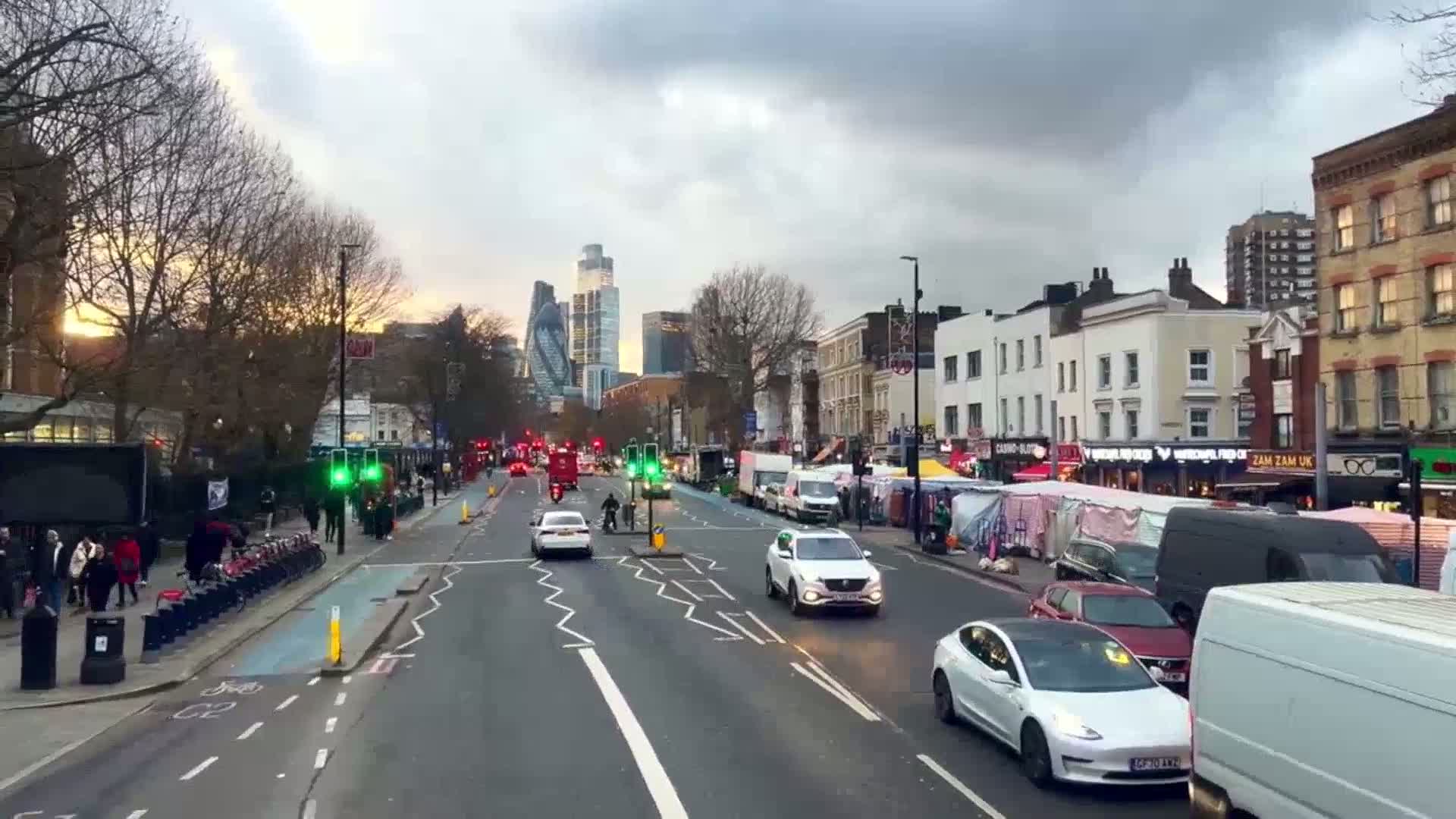 A city street scene at sunset under partly cloudy skies shows several cars and vans driving past market stalls and buildings, with tall skyscrapers visible in the distance.