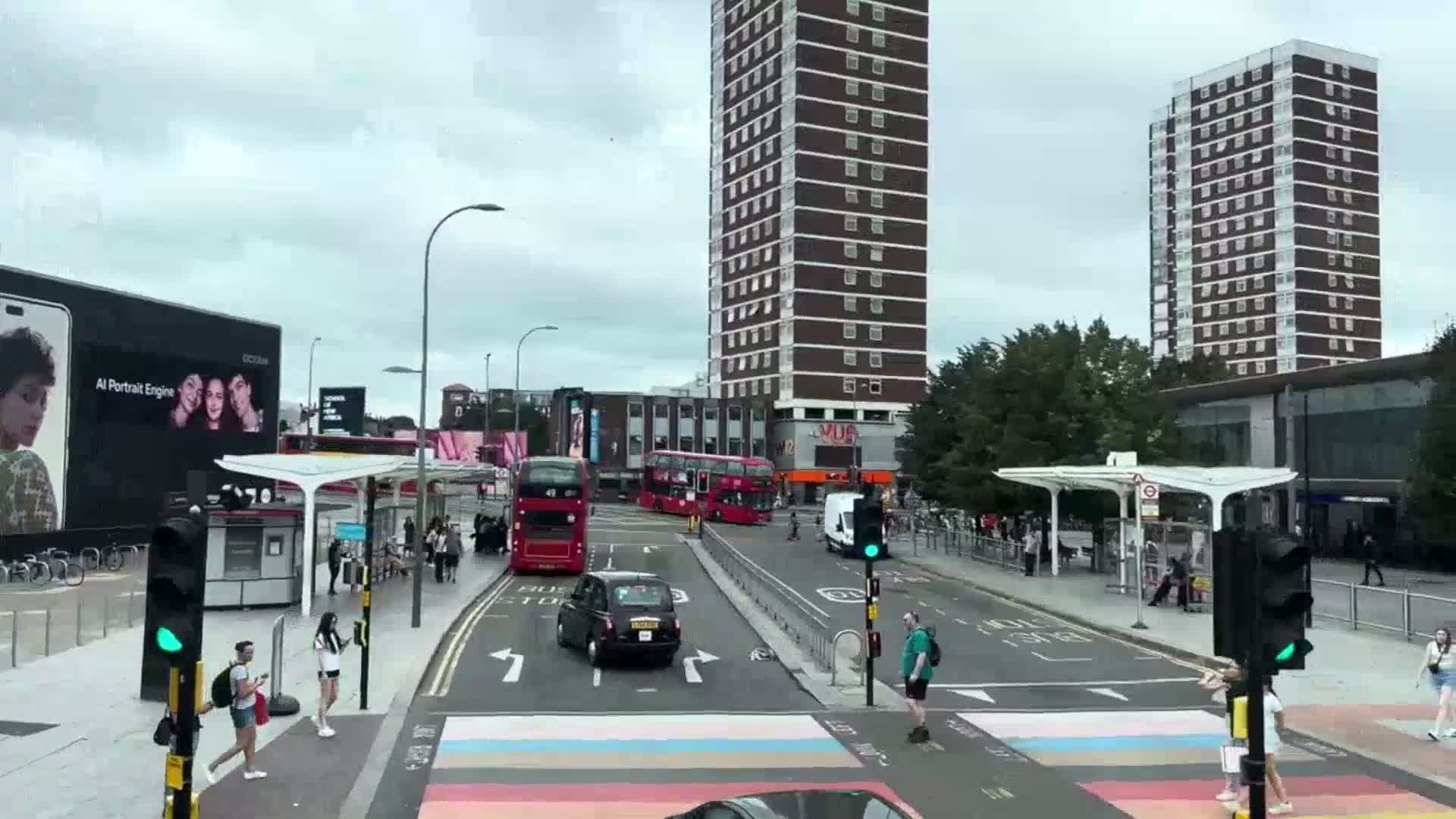 A city street scene on an overcast day shows several buses and a taxi at an intersection near tall apartment buildings and bus shelters with pedestrians crossing a rainbow crosswalk.
