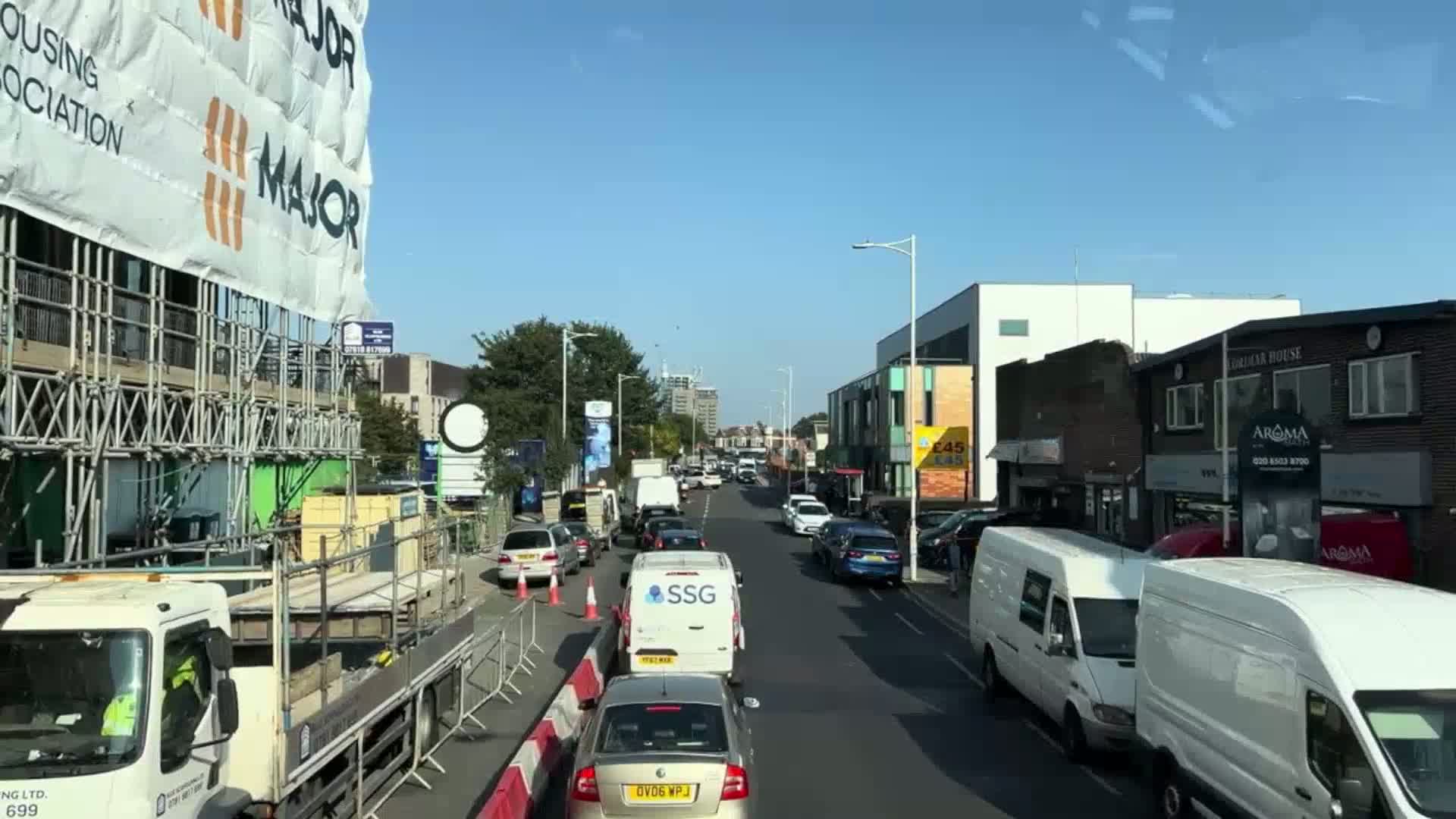 A sunny day reveals a busy street scene with construction on one side and shops and parked vans on the other, with traffic flowing in both directions.